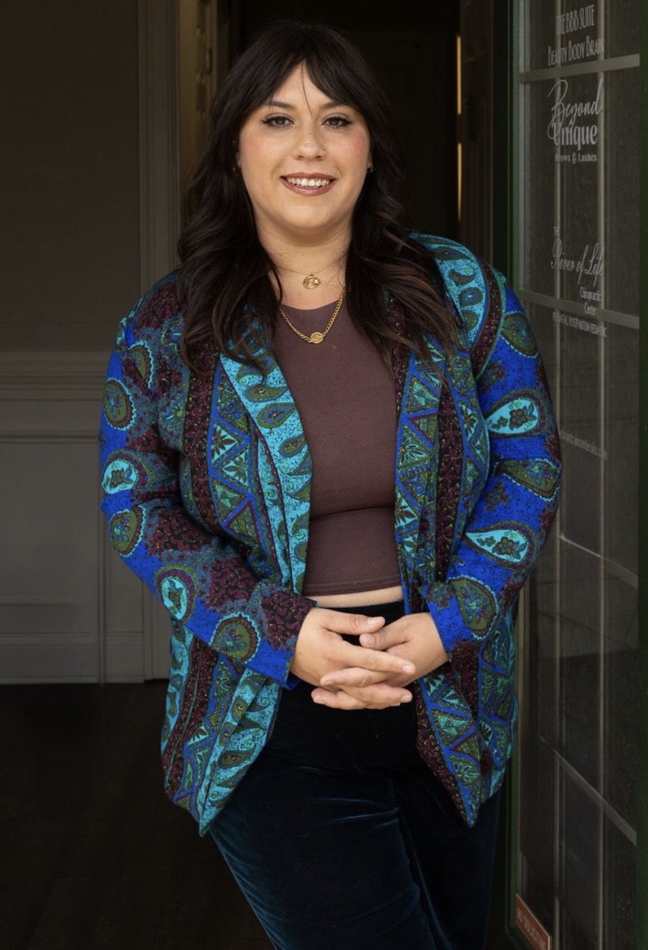 Woman with dark hair smiling, wearing a patterned blue jacket and dark top, standing indoors near a chalkboard.