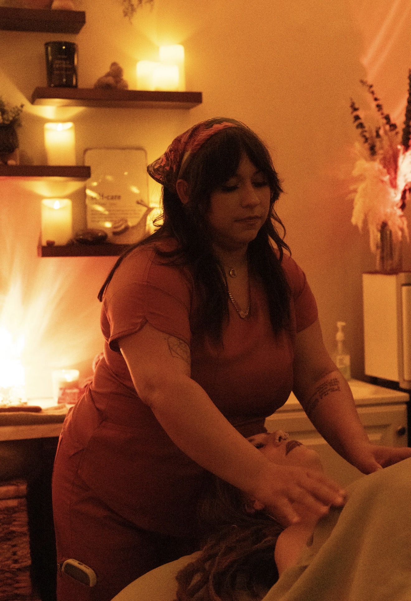 Woman with dark hair and a headscarf in a warm-lit room, touching a person's head, with shelves and flowers in background.