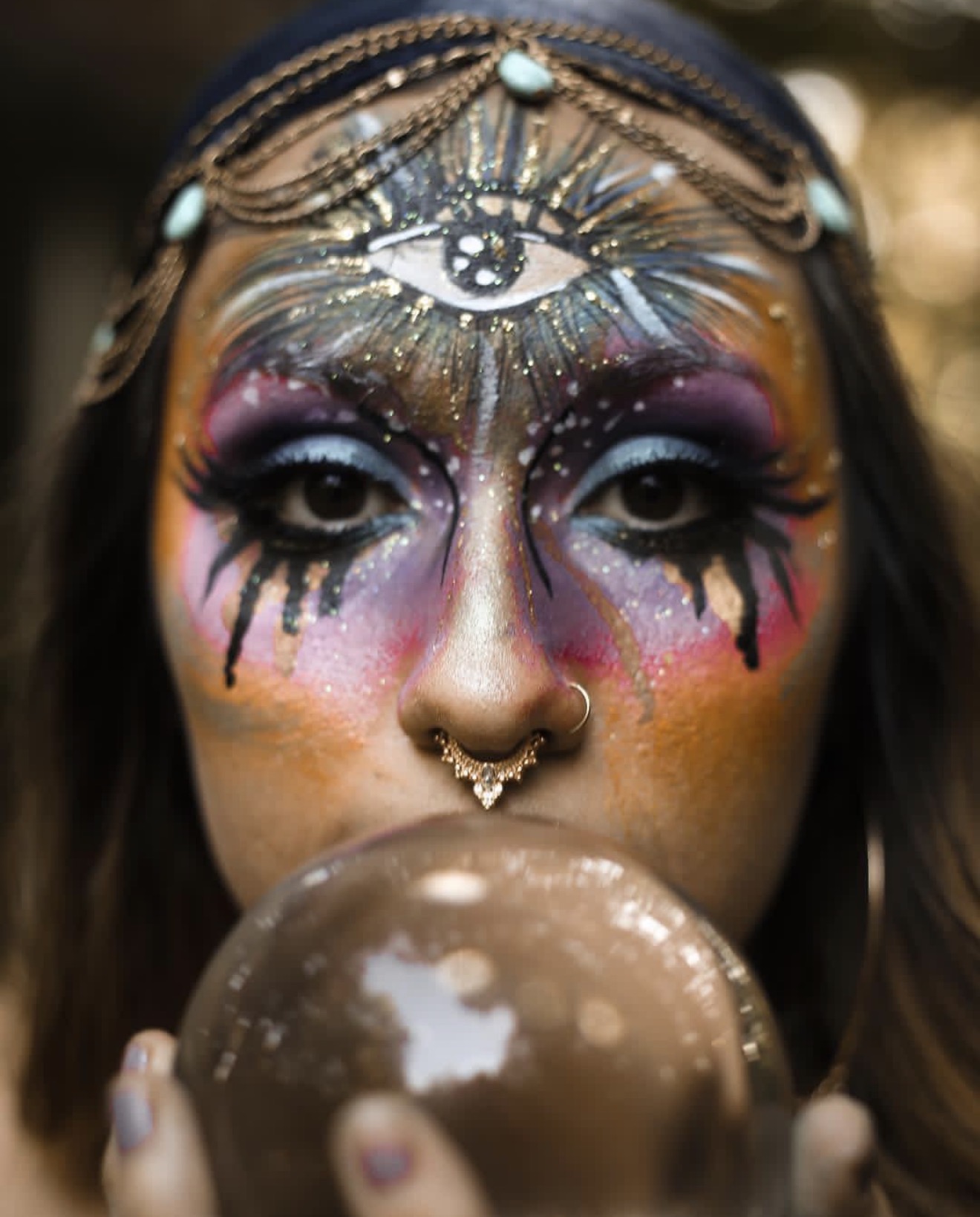 Person with elaborate face makeup, including an eye design on forehead, colorful eyeshadow, and holding a reflective sphere.