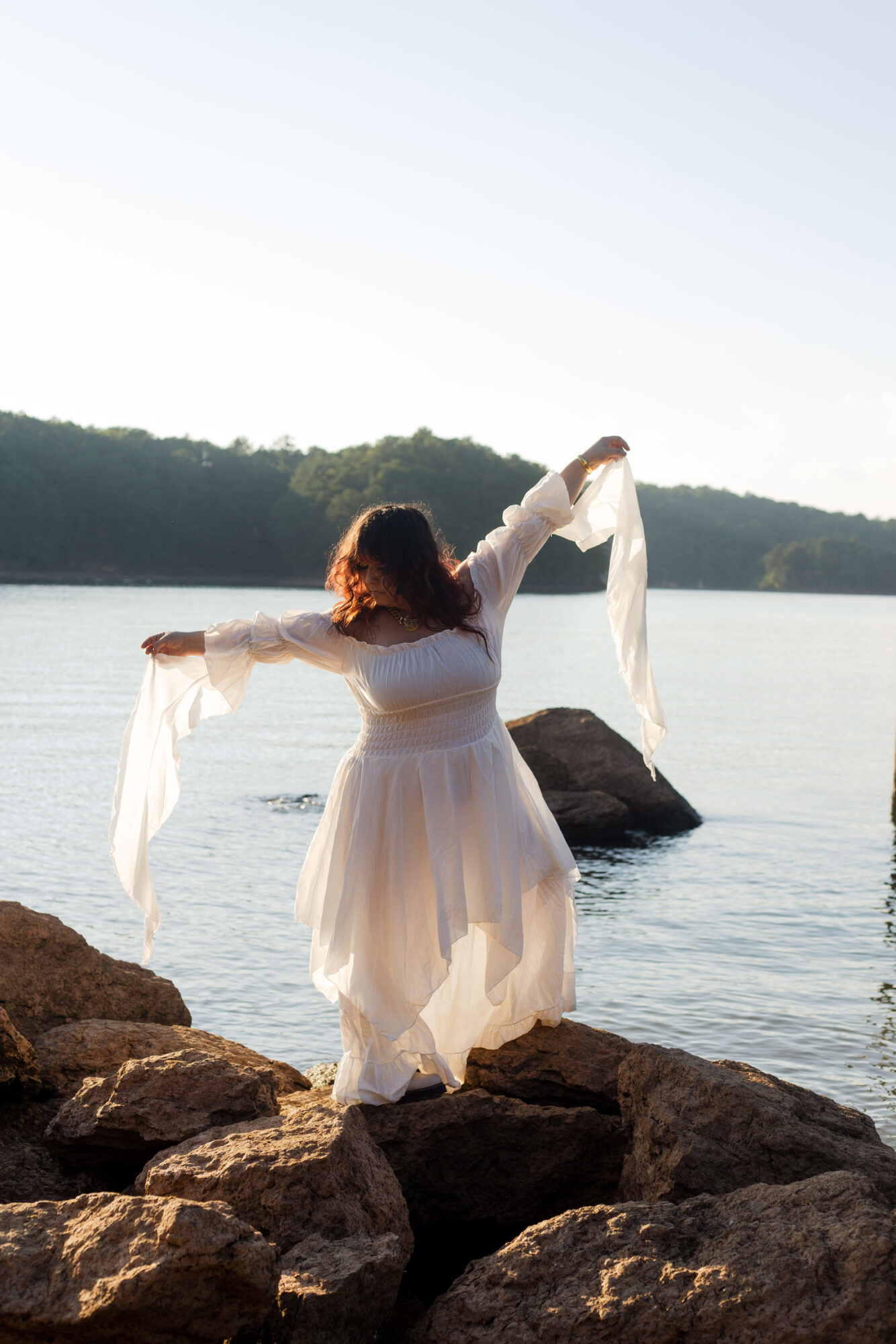 Person in white dress standing on rocks by water with arms outstretched, facing away, during daytime.