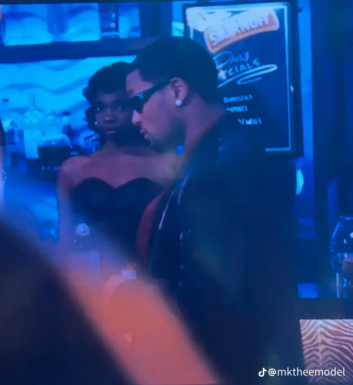 Two people in a dimly lit setting, one wearing glasses and a dark outfit, the other in a strapless top, with a neon sign in the background.