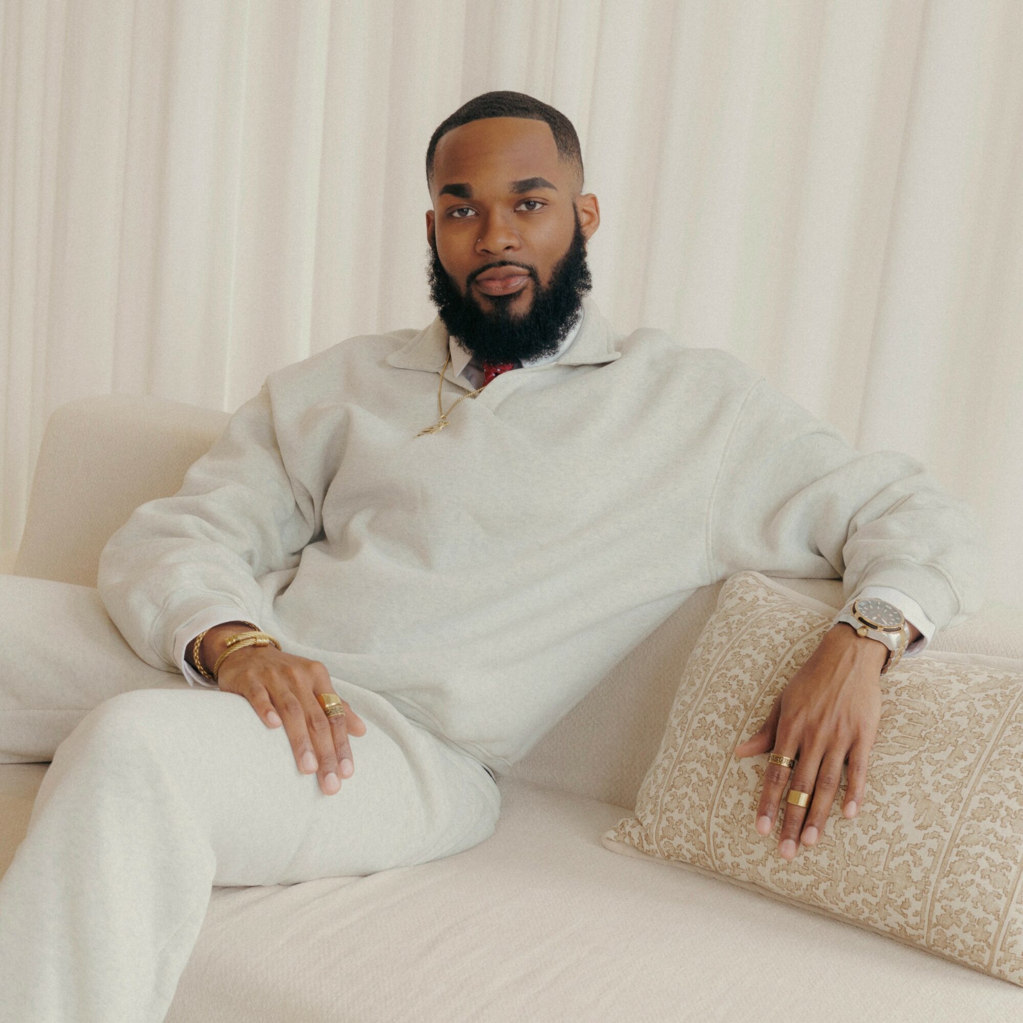 Man with a beard sitting on a sofa, wearing a light-colored sweater and pants, with a curtain background.