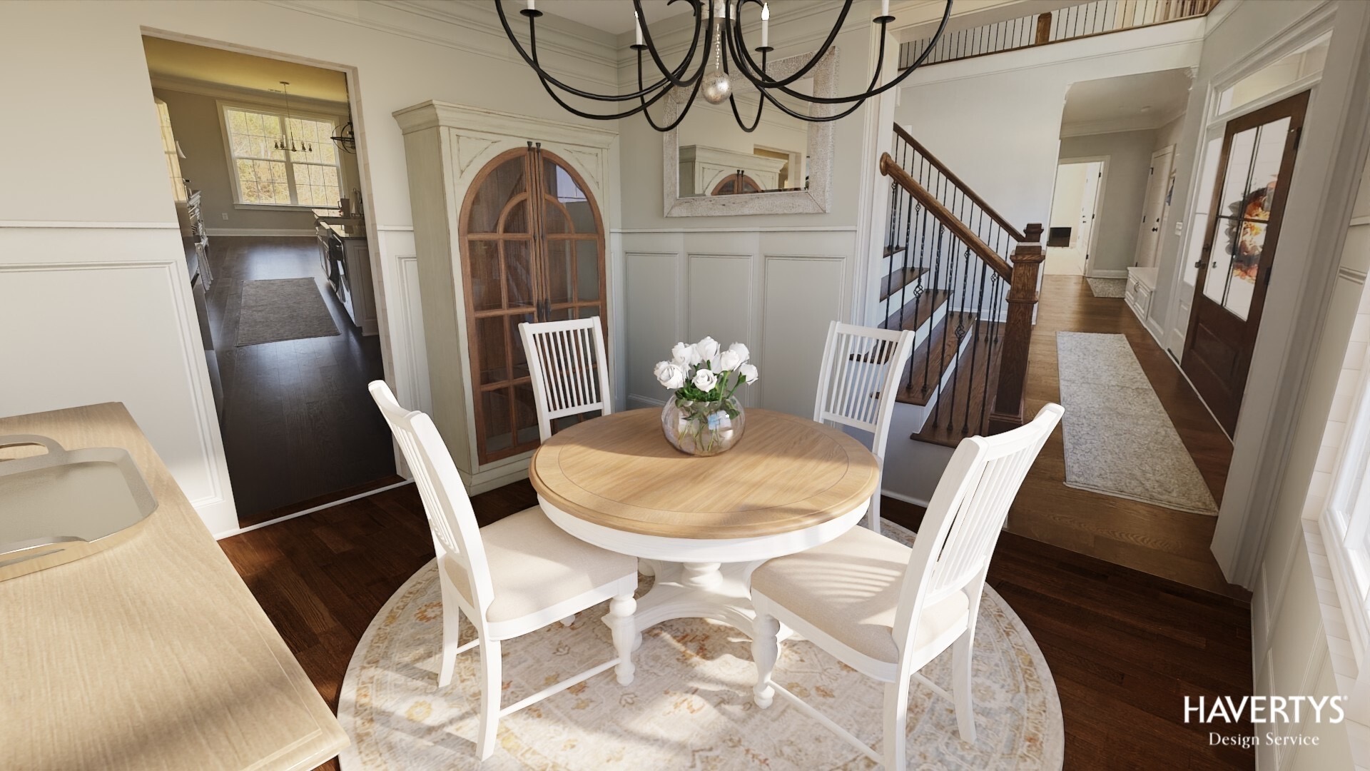 Dining area with round wooden table, six white chairs, a floral centerpiece, and a chandelier, in a bright room with stairs.