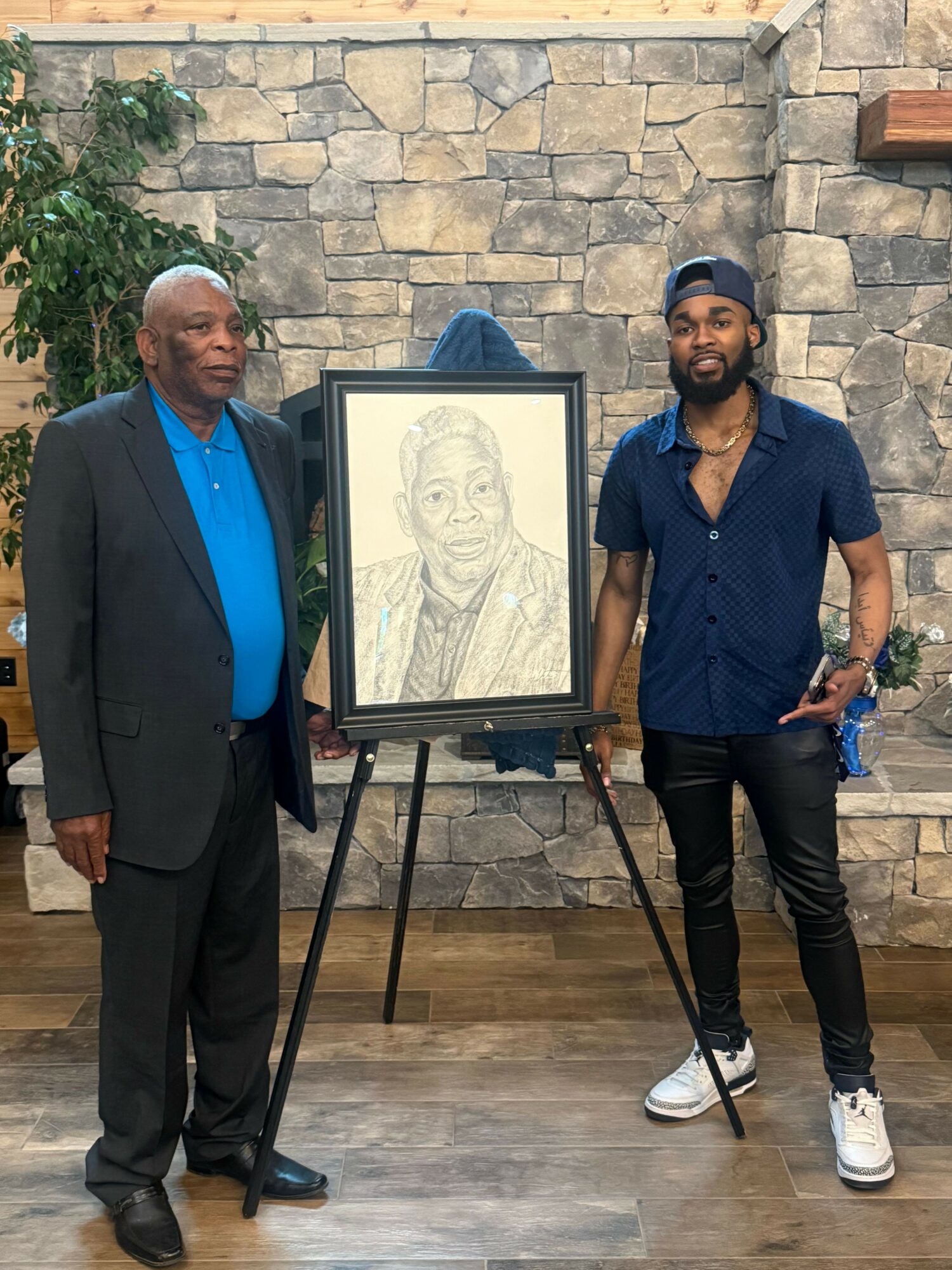 Two men standing indoors in front of a stone wall, with a framed portrait on an easel between them.
