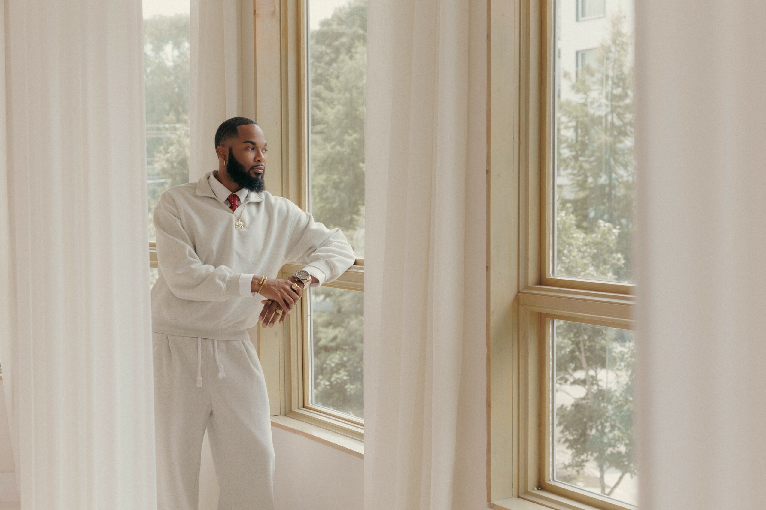 Man standing by large window with curtains, looking outside, with trees visible through the window.