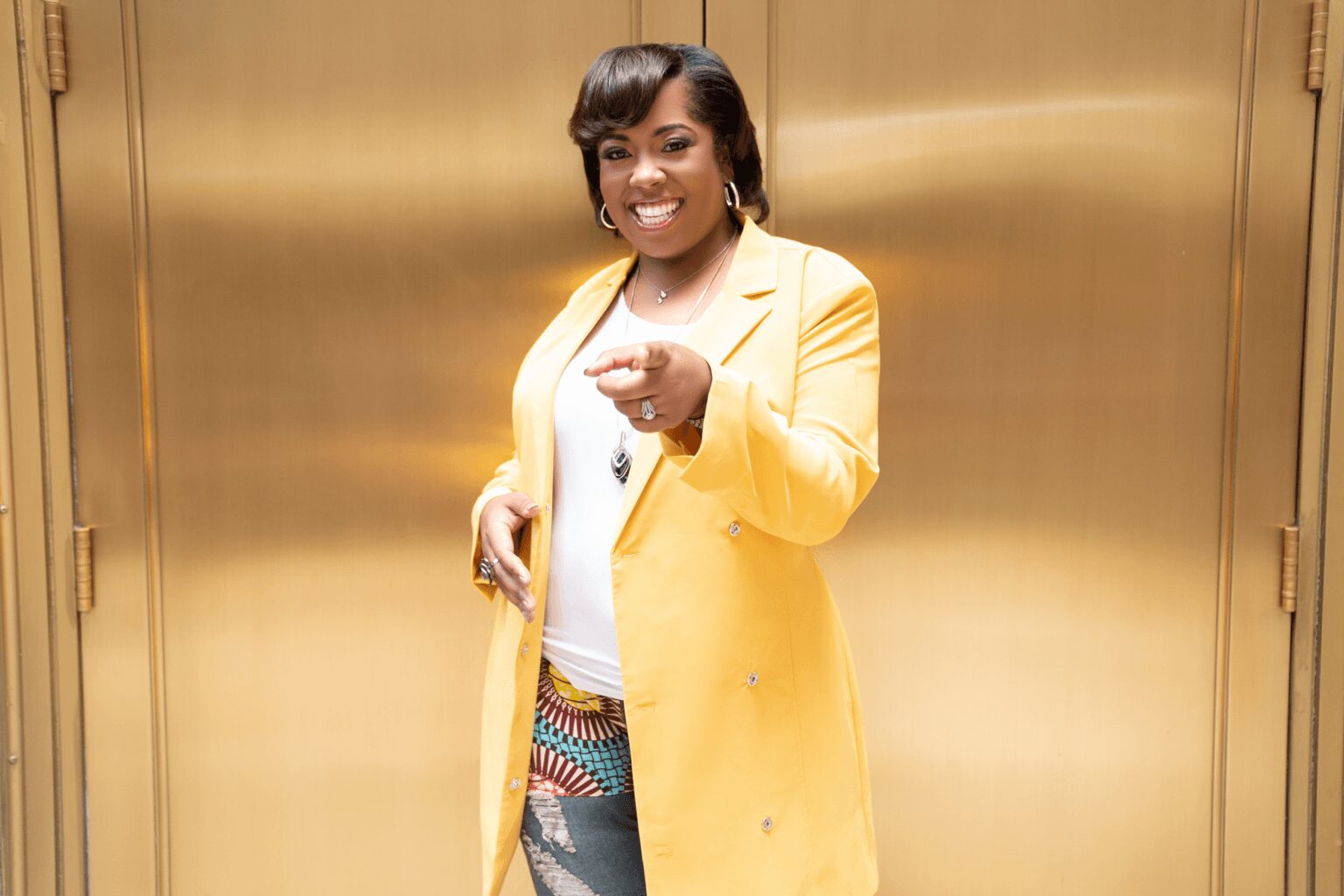 Woman in yellow blazer smiling and pointing at camera, standing in front of a gold background.
