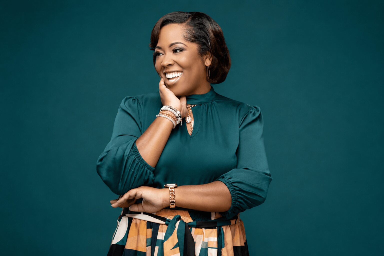 Smiling woman with short hair wearing a teal blouse and patterned skirt, posing with her hand near her face, against a teal background.