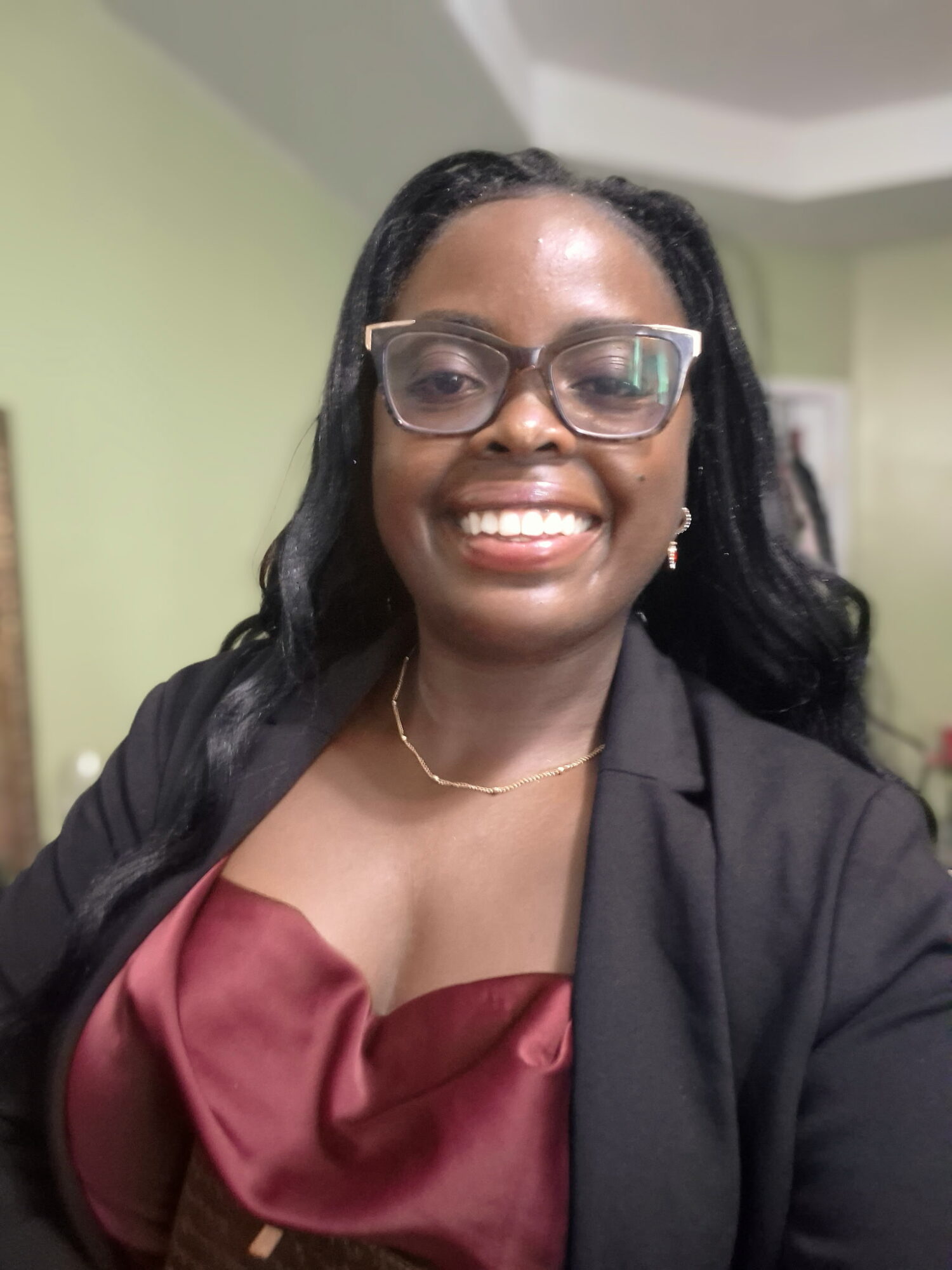 Smiling woman with glasses, wearing a black blazer and maroon top, in an indoor setting.