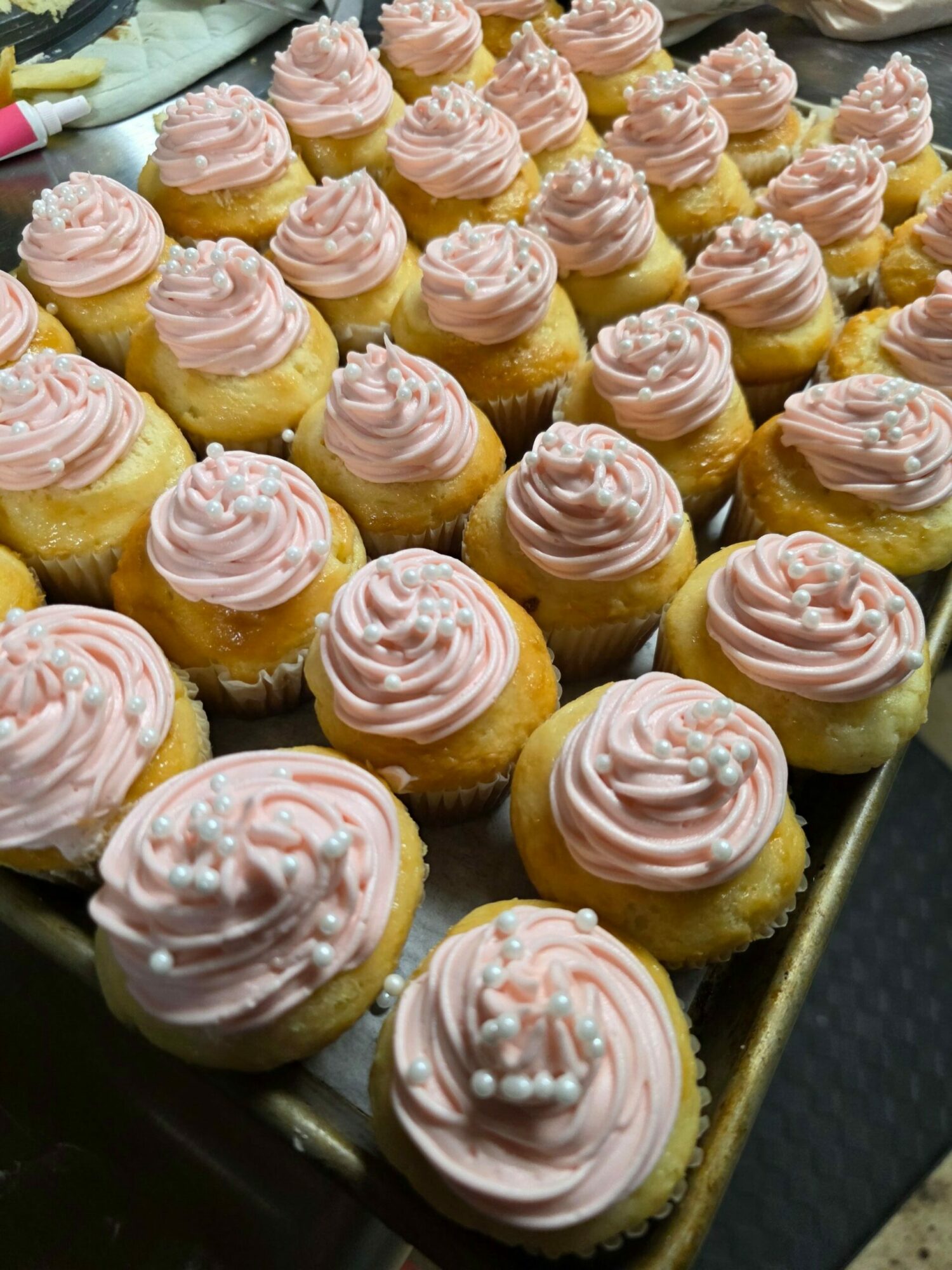 Tray of cupcakes with pink frosting and sprinkles, arranged in rows, viewed from above.