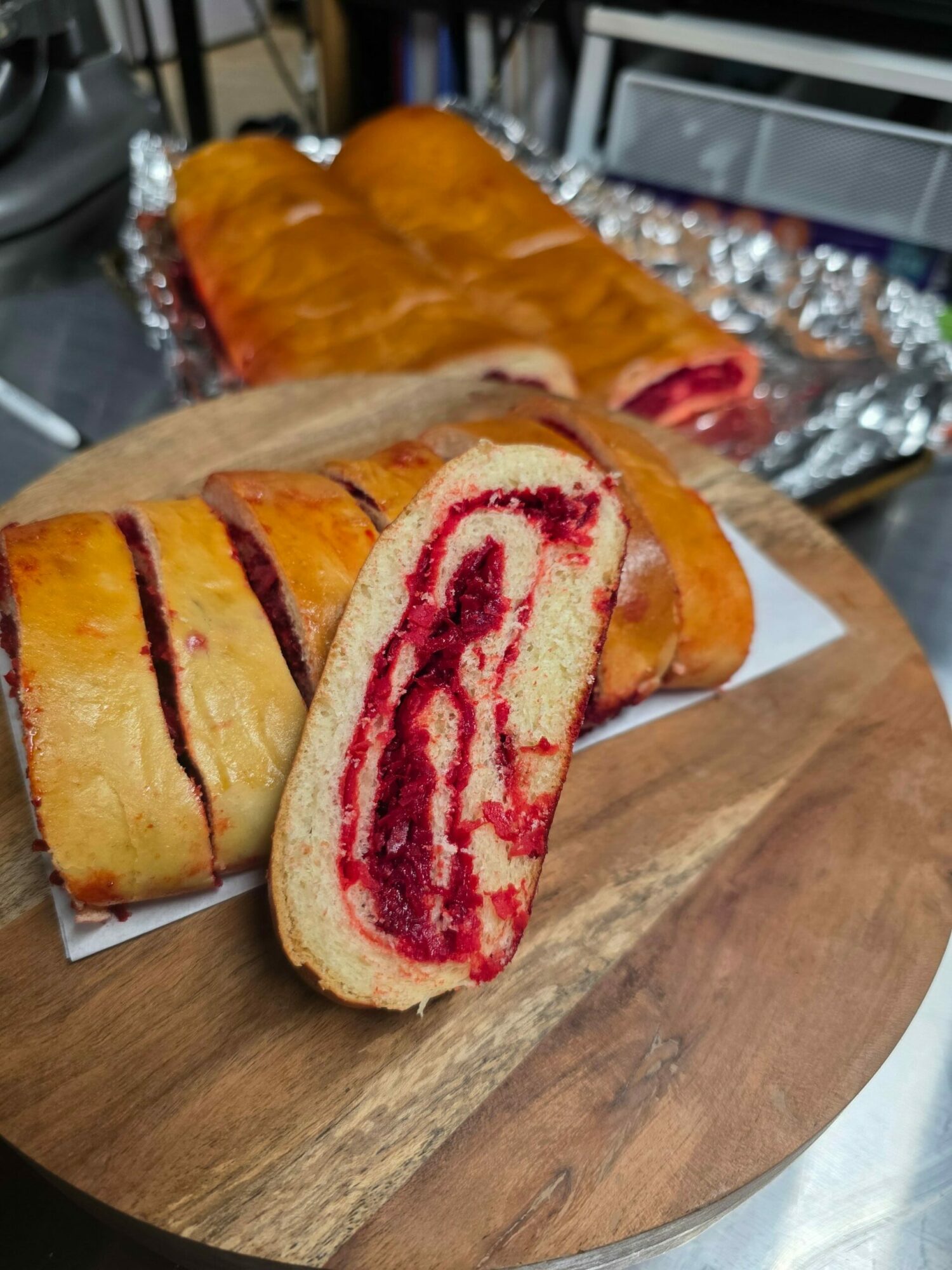 Sliced pastry with red filling on a wooden cutting board, with a whole pastry in the background, on a white paper sheet.