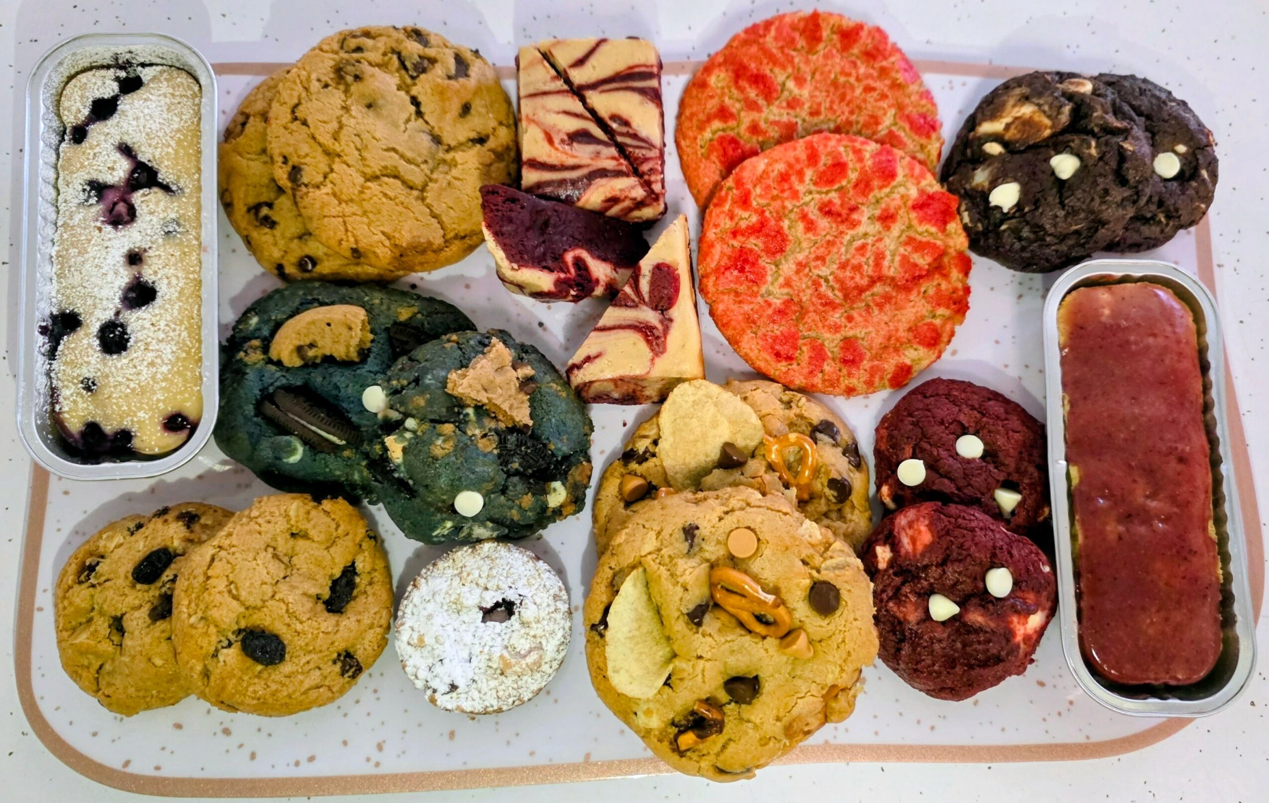 Assorted cookies and baked goods on a tray, including chocolate chip, fruit, and decorated cookies, with a rectangular dessert on the right.
