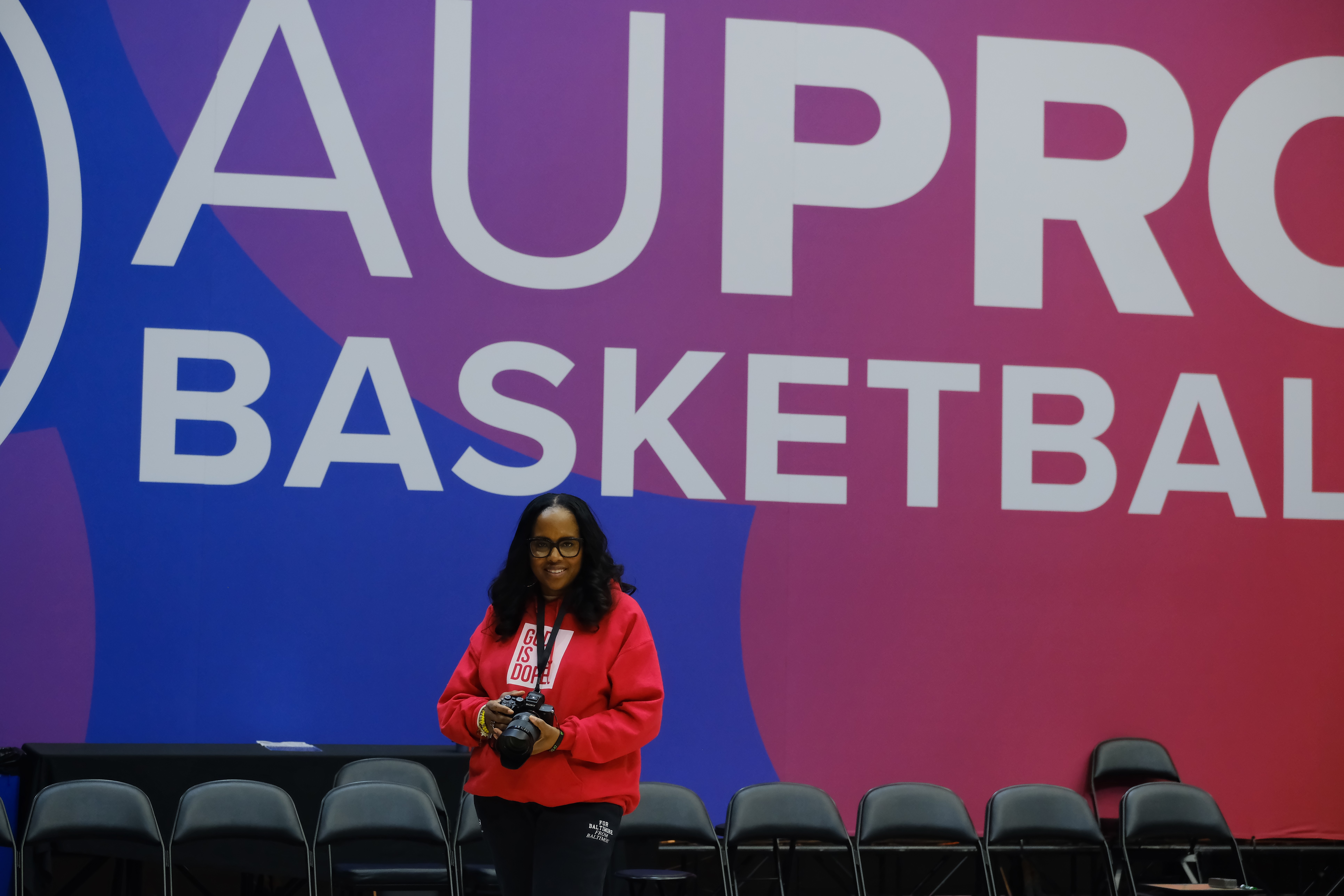 Woman in red jacket standing in front of a large purple and pink sign that reads 'AUPRO BASKETBALL'.