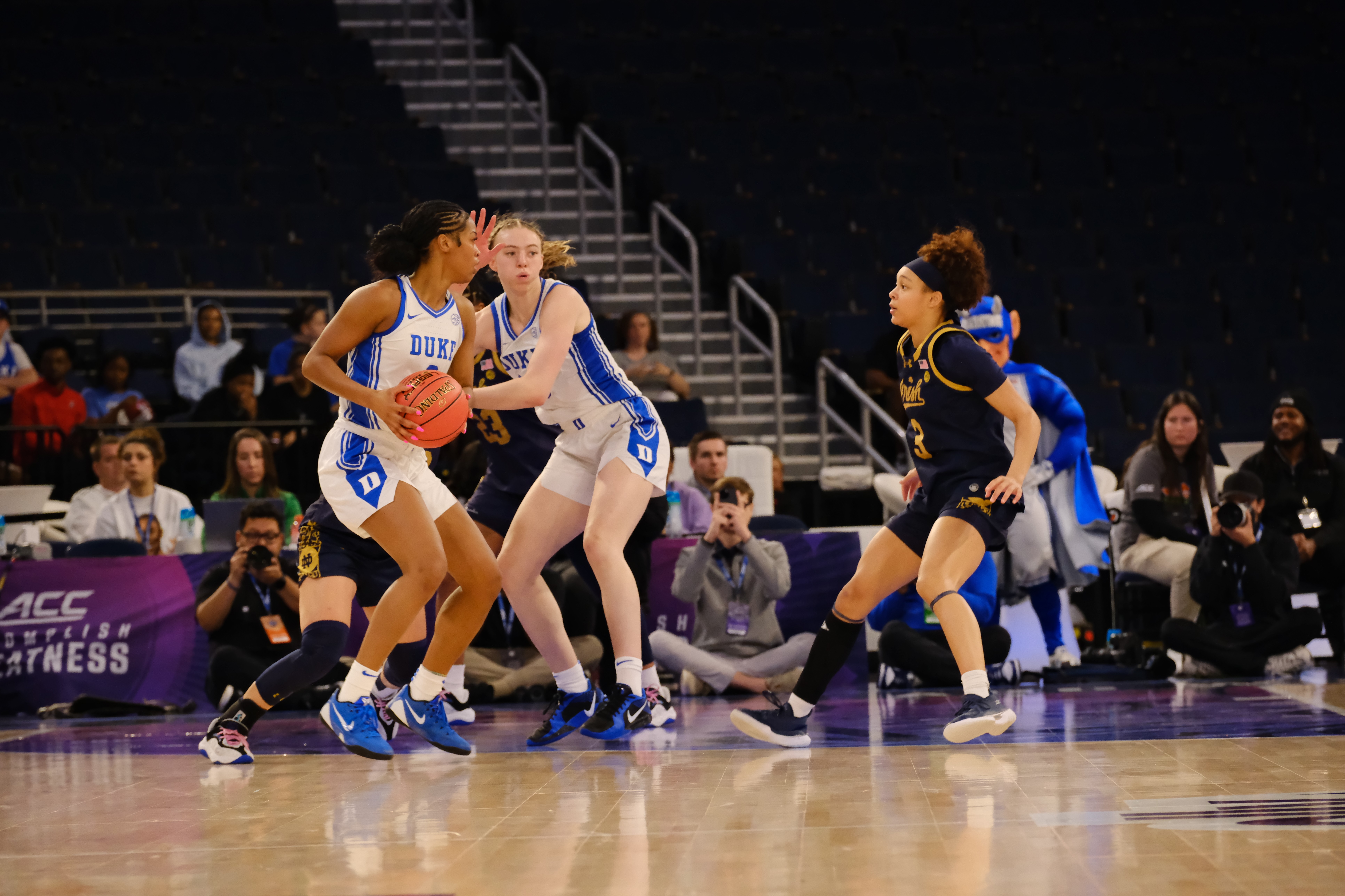 Four female basketball players on court, two in white jerseys and two in dark jerseys, with spectators in background.