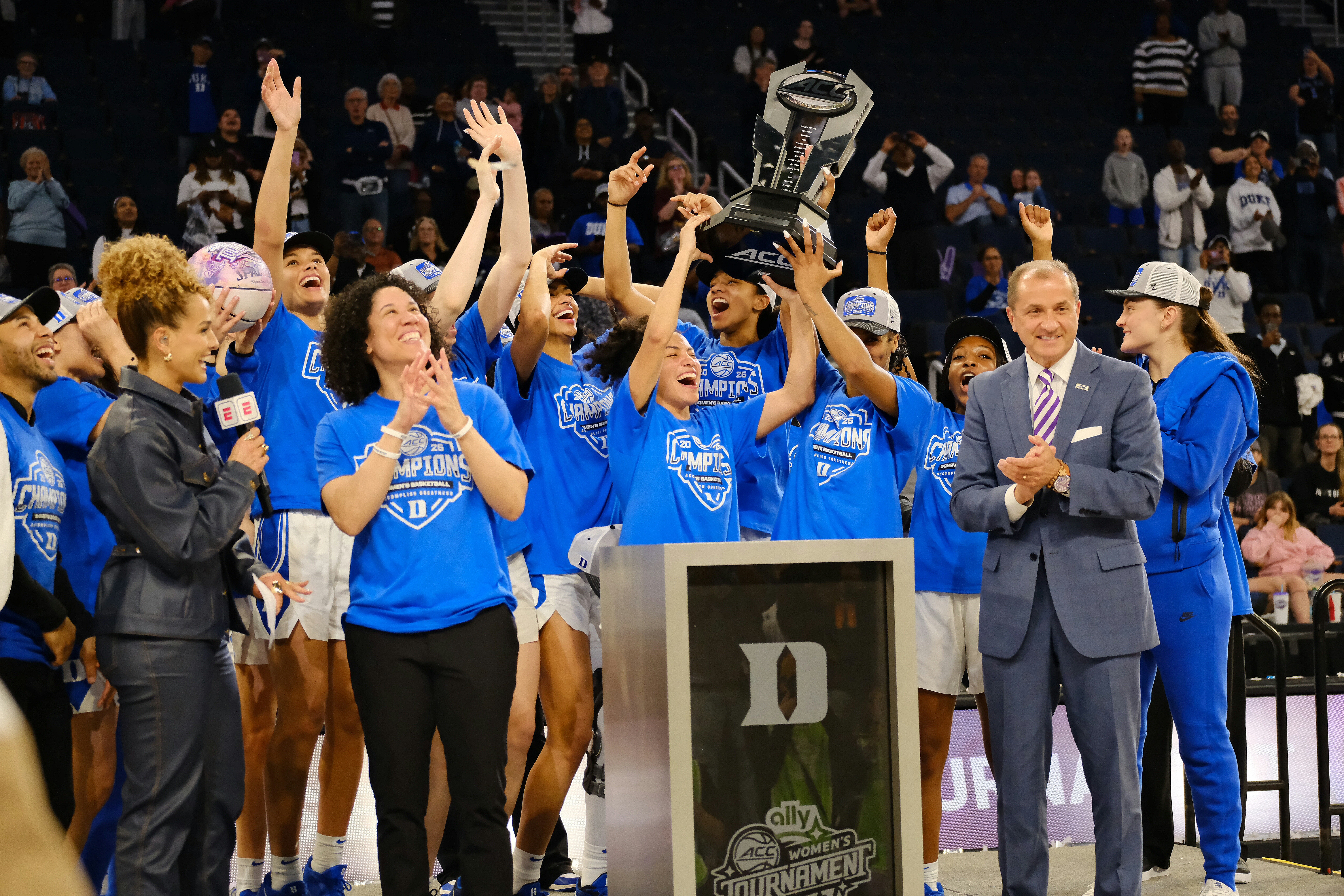Group of people celebrating with raised hands, wearing blue shirts, and a man in a suit clapping, in an indoor sports arena.