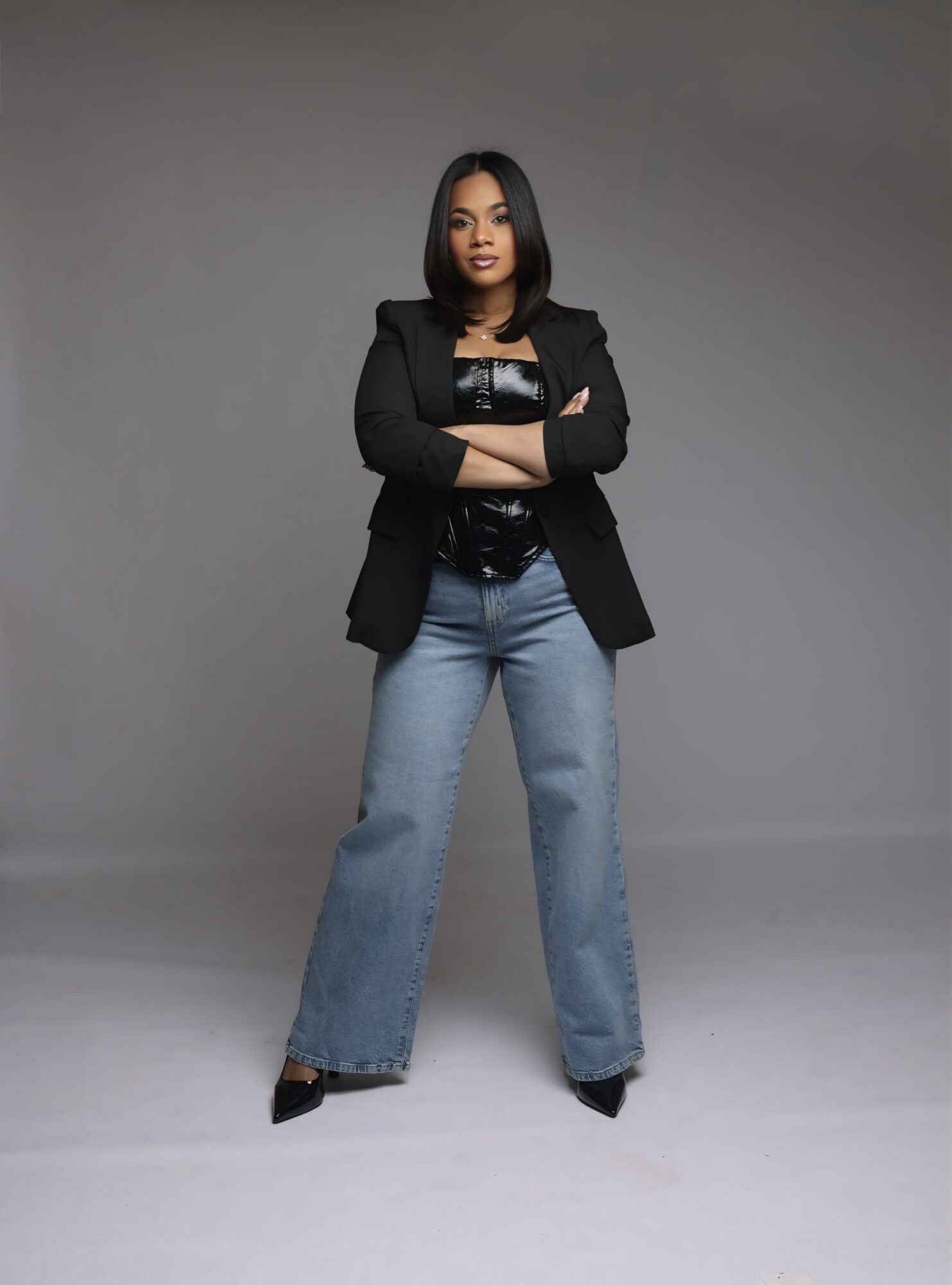 Woman standing with arms crossed, wearing a black blazer, black top, and wide-leg jeans, against a gray background.