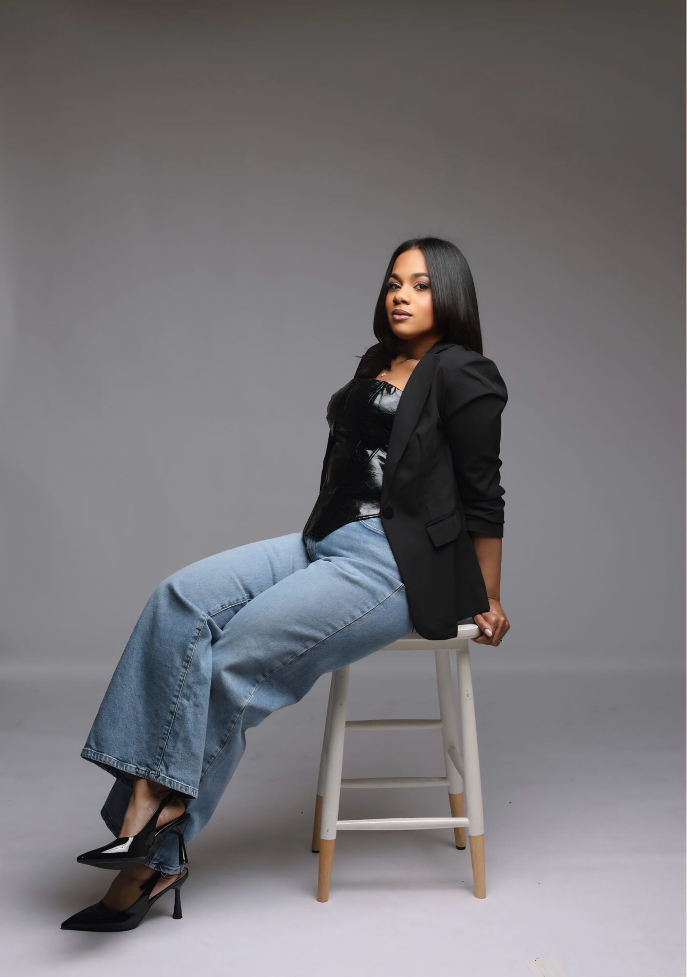 Woman sitting on a stool, wearing a black blazer, black top, light blue wide-leg jeans, and black high heels, against a plain gray background.