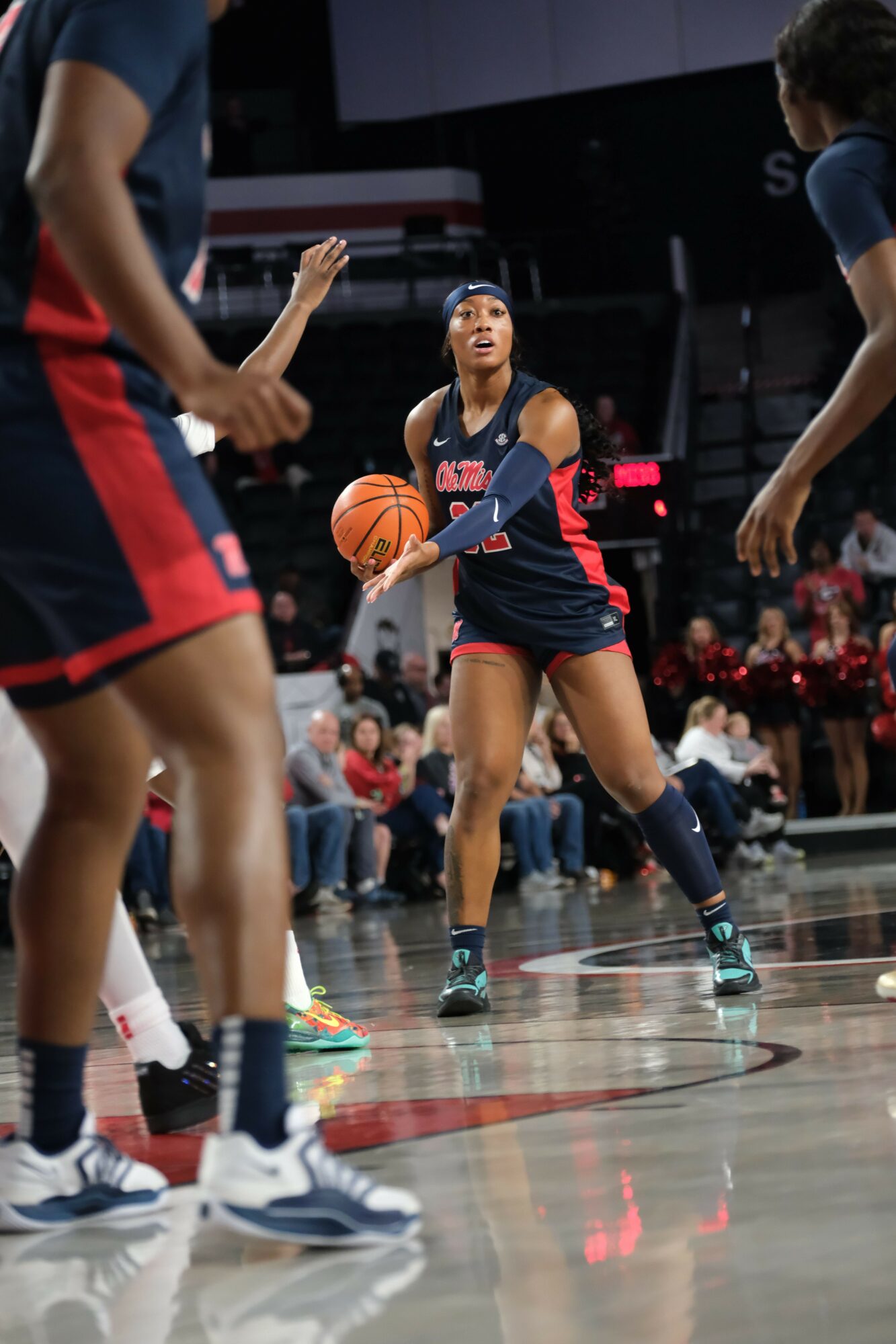 Female basketball player in a dark uniform dribbling a basketball on court, with other players and spectators nearby.