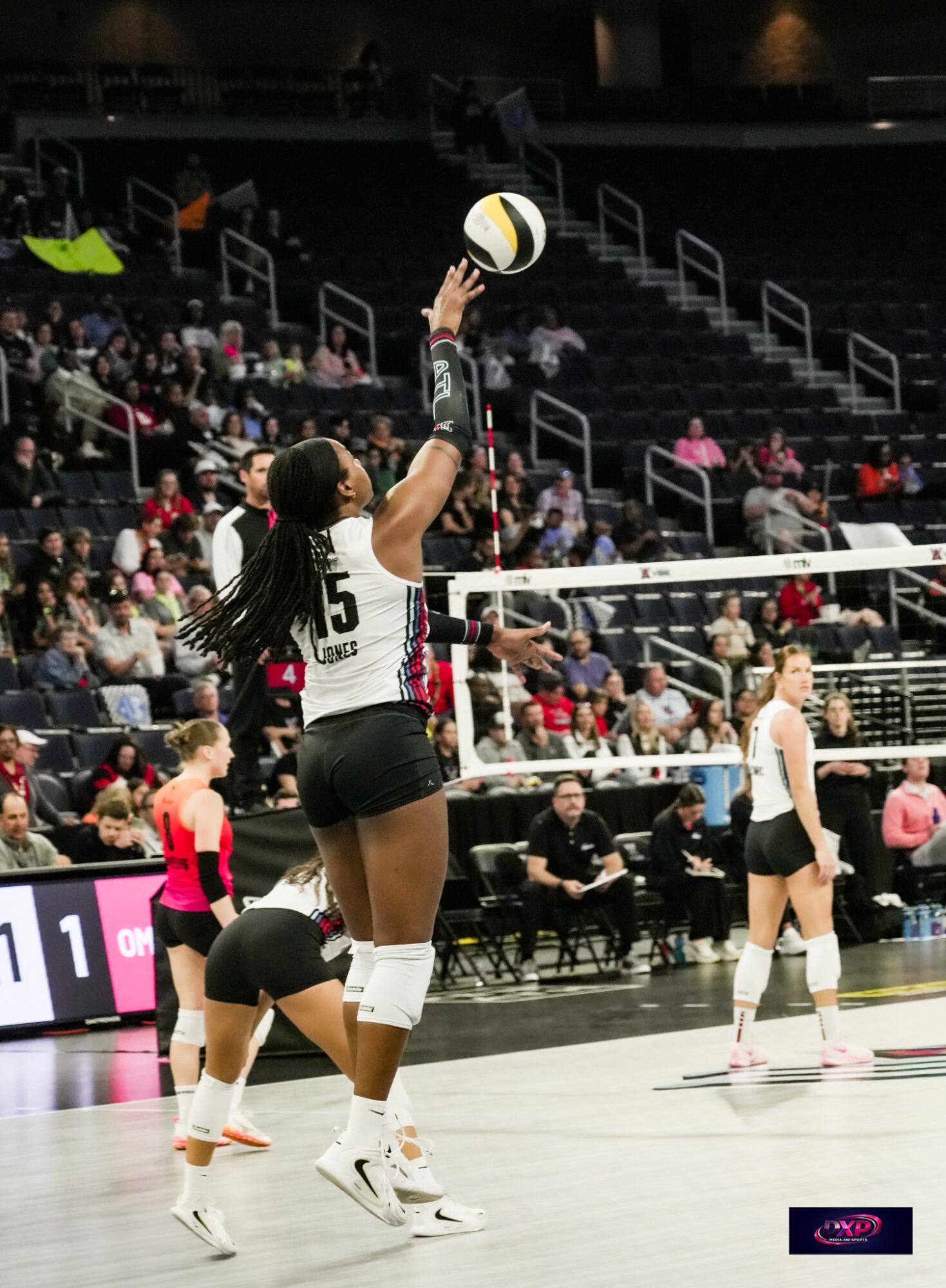 Female volleyball player in black and white uniform jumps to hit the ball during a match, with spectators in the background.