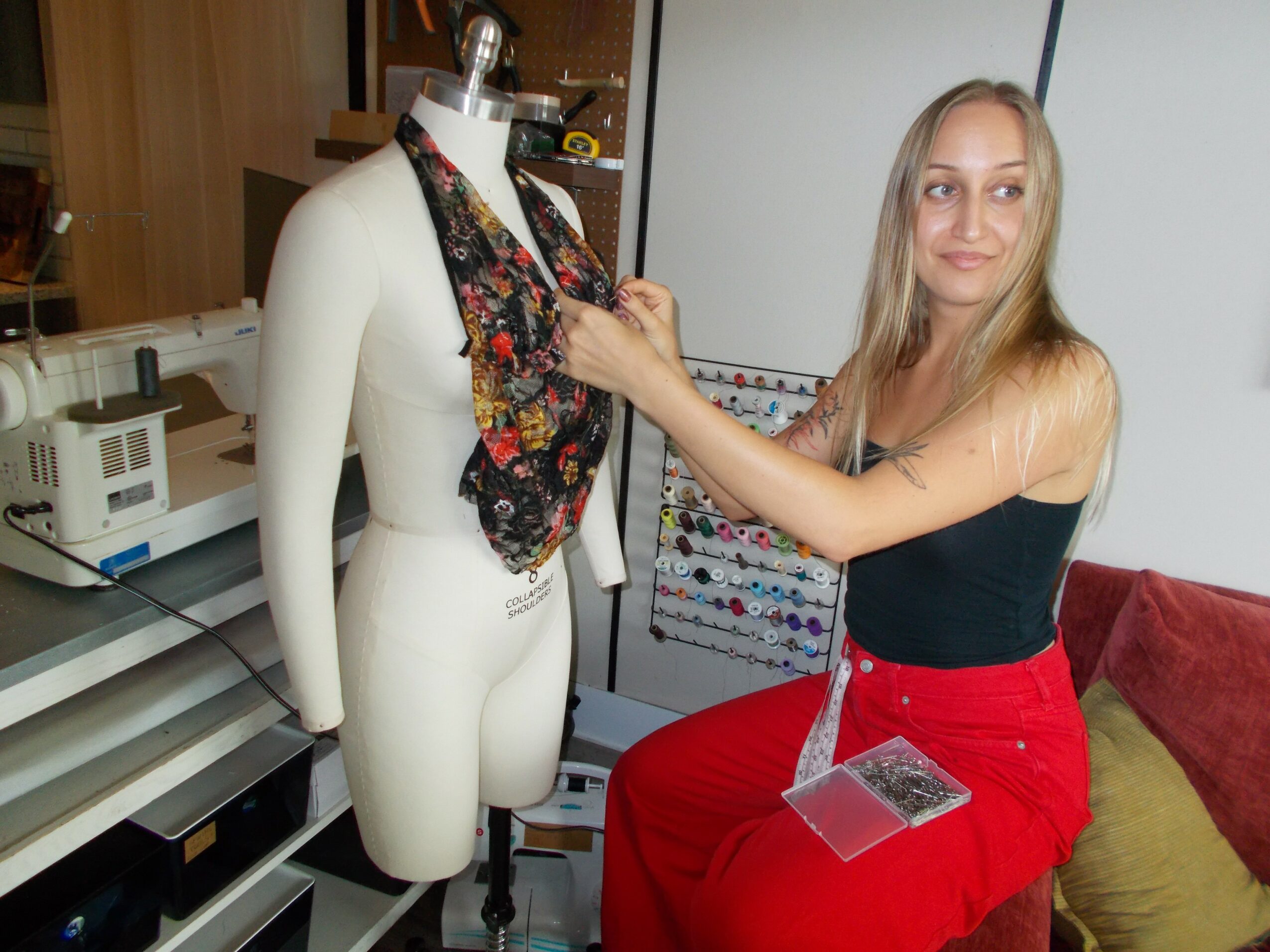 Woman with long blonde hair, black top, and red skirt, smiling and touching a mannequin with a floral scarf.