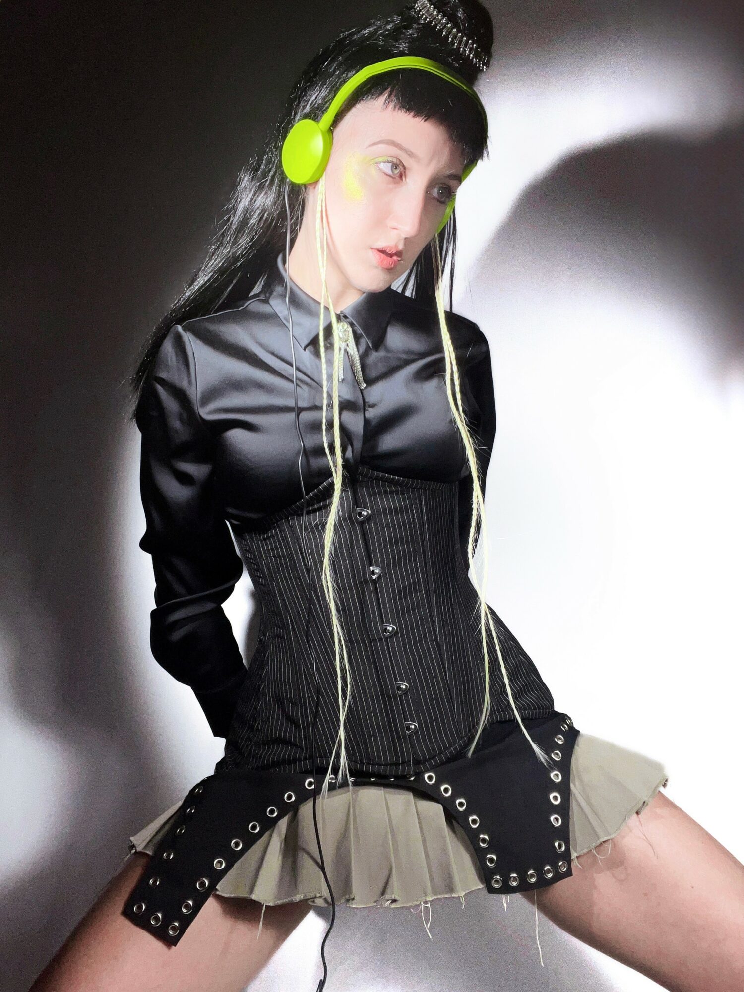 Young woman with black hair and green headphones, wearing a black jacket and beige skirt, sitting against a wall.