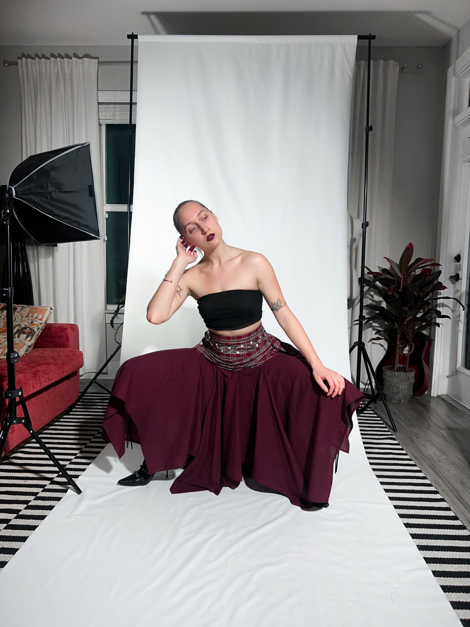 Person sitting on the floor in front of a white backdrop, wearing a black top and a long maroon skirt, smiling.