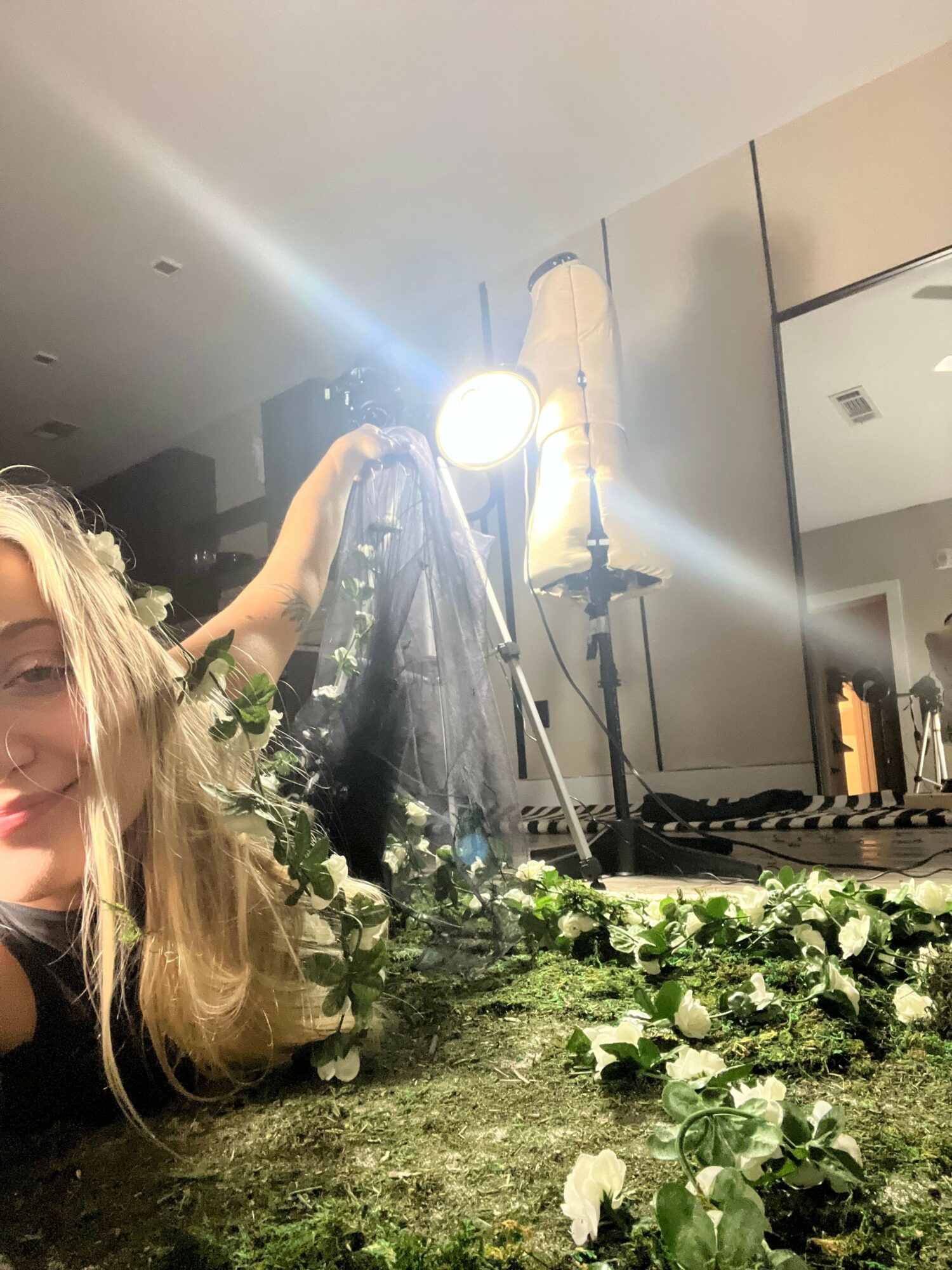 Person with long hair working with plants and flowers indoors, with a bright light and kitchen in background.