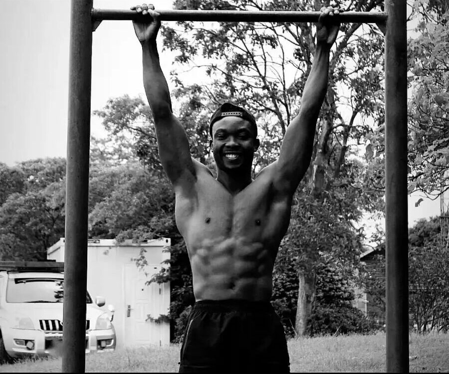 A shirtless man with glasses hangs from a horizontal bar outdoors, smiling, with trees and a vehicle in the background.
