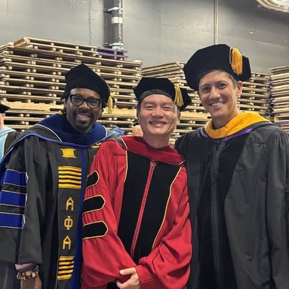 Three graduates in caps and gowns smiling, standing in front of stacked wooden pallets, celebrating graduation.