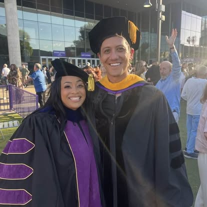 Two graduates in caps and gowns smiling outdoors with a crowd and modern building in background.