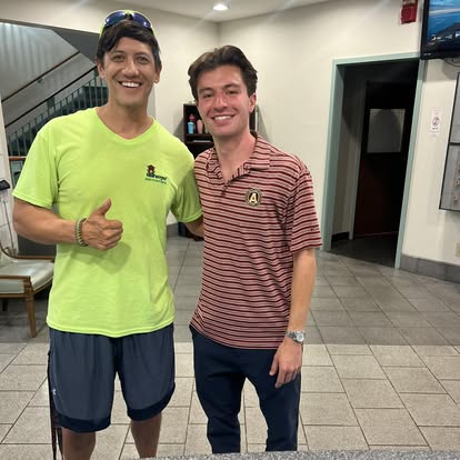 Two men standing indoors, smiling, one giving a thumbs-up, in casual clothing, with a staircase and door in the background.