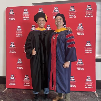 Two graduates in academic regalia standing in front of a red backdrop with university logos, smiling and posing for a photo.