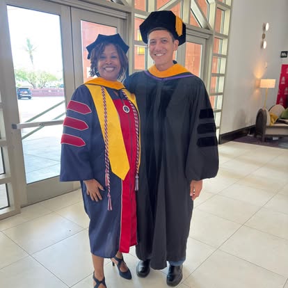 Two graduates in academic regalia standing indoors, smiling, with large windows and modern interior in background.