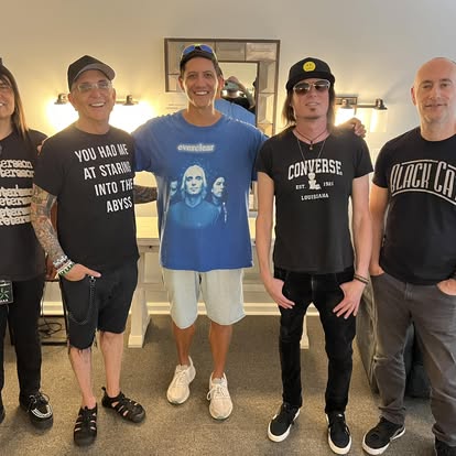 Group of five people standing indoors, smiling, with a mirror and lights in the background.
