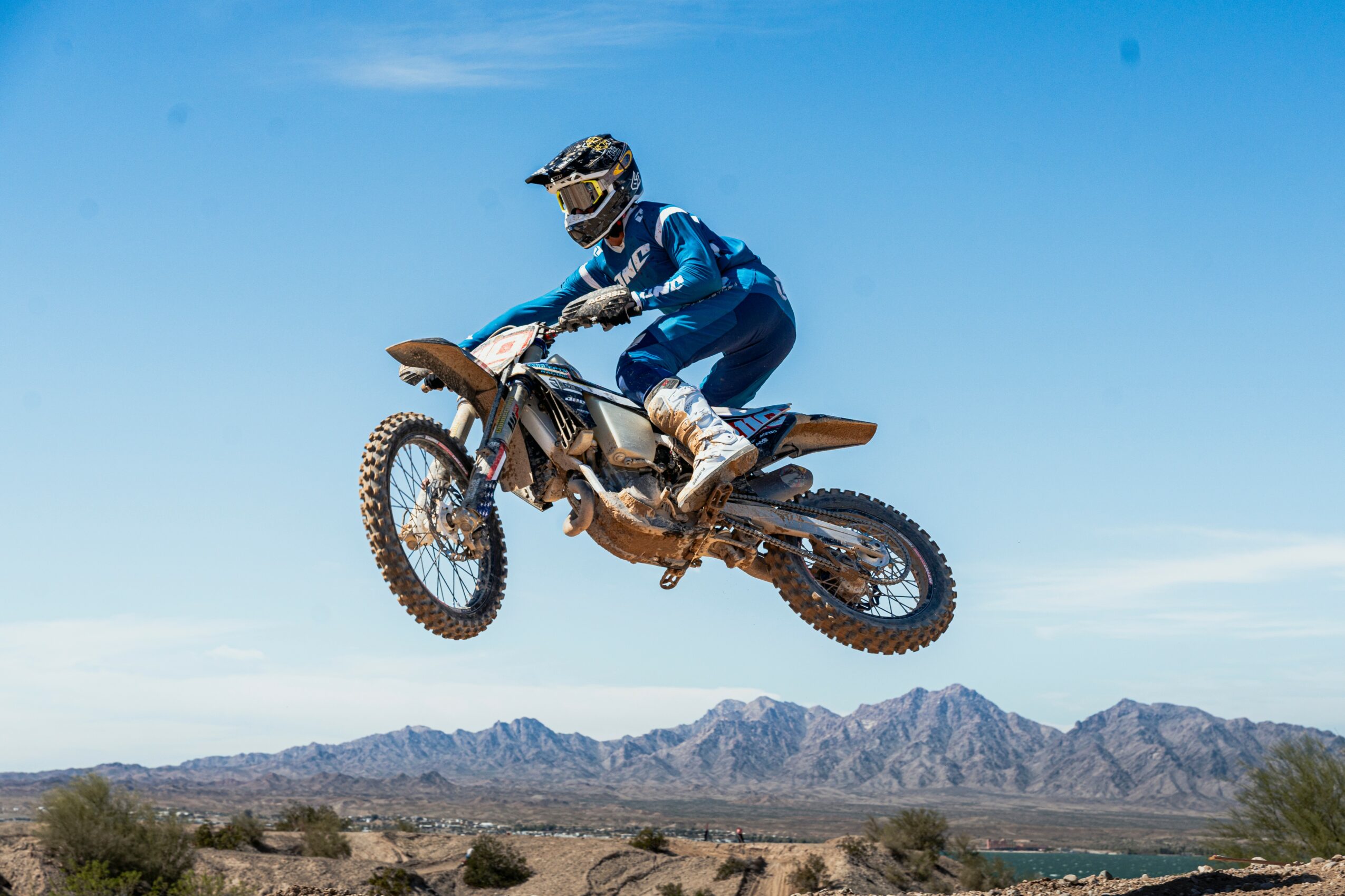 Person in blue gear riding a dirt bike mid-air over desert landscape with mountains in background