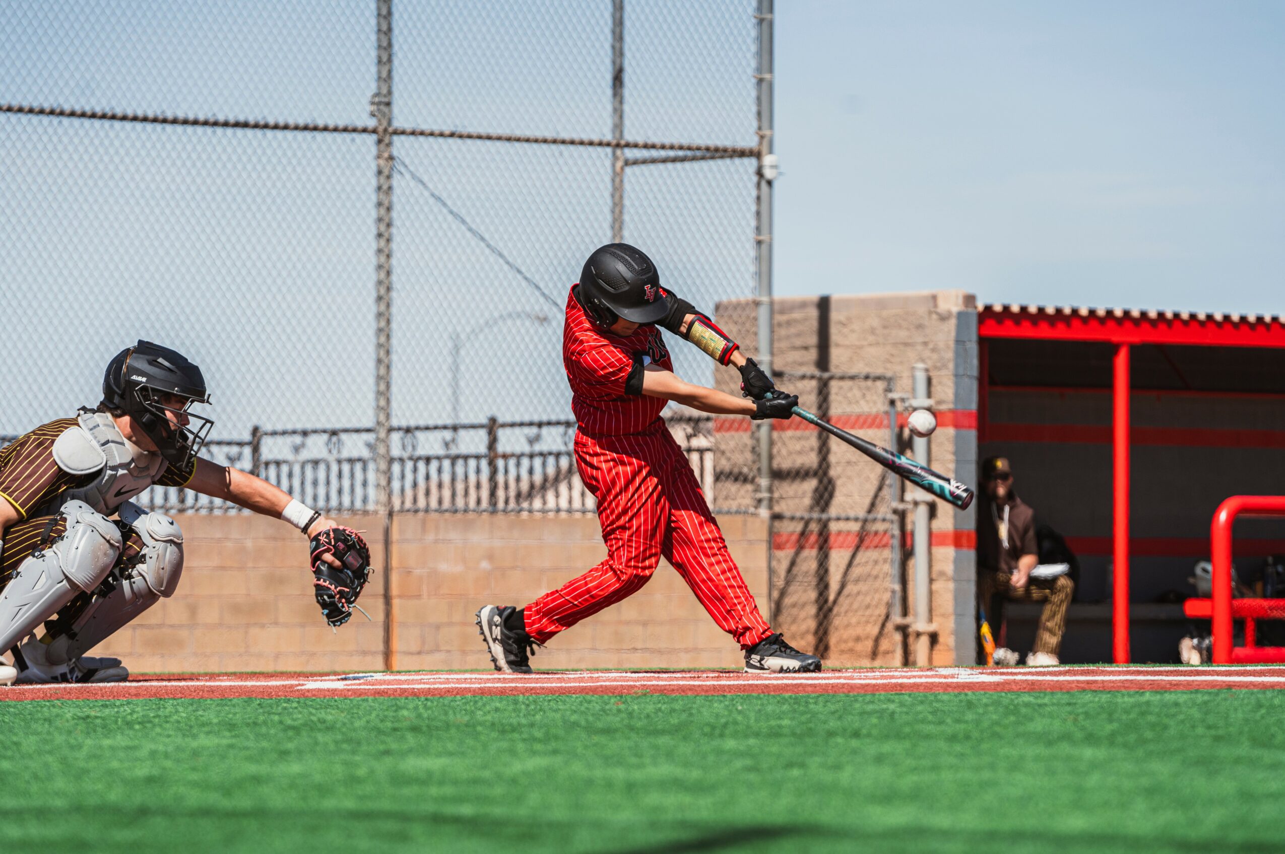 Baseball player swings bat at ball, catcher crouches behind, field and dugout in background.