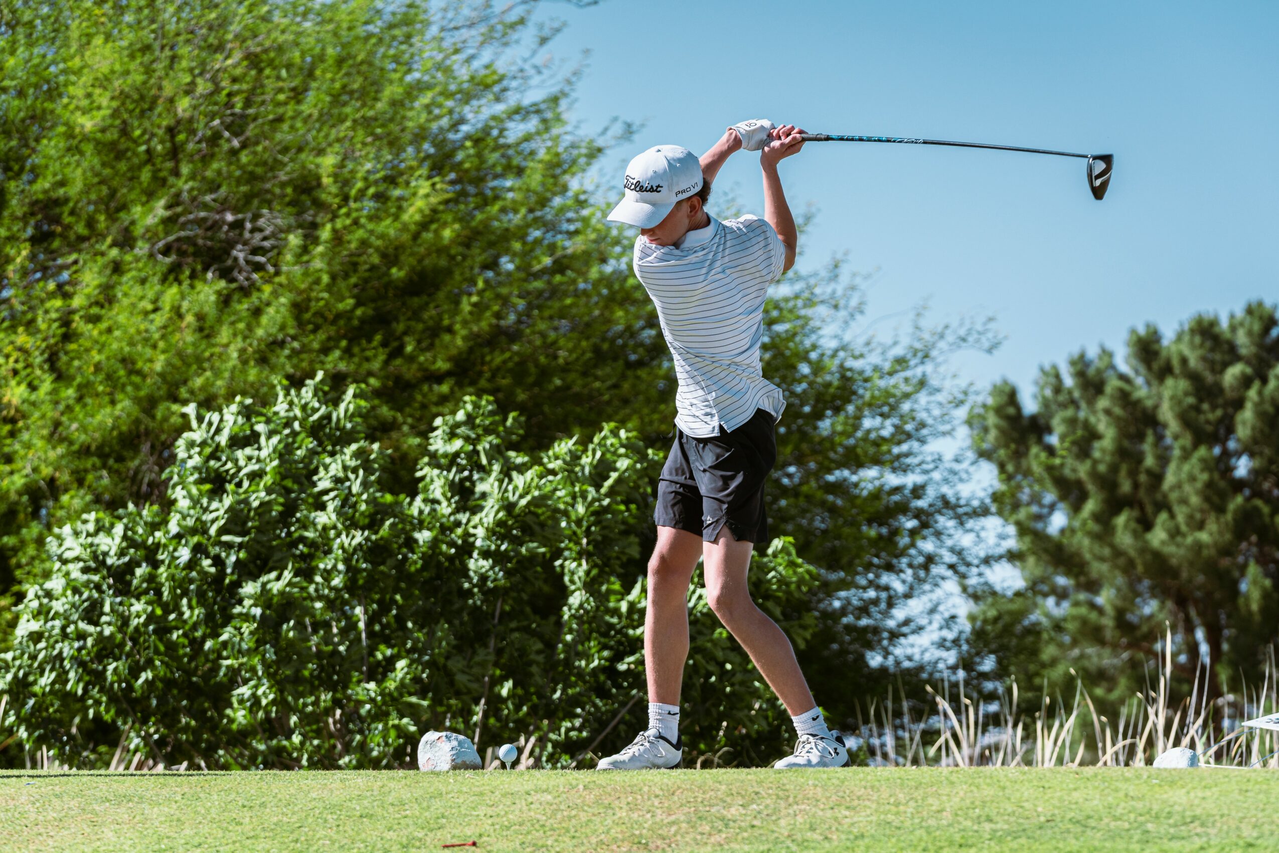 Child in striped shirt and hat swings a golf club on a sunny day with trees in the background.