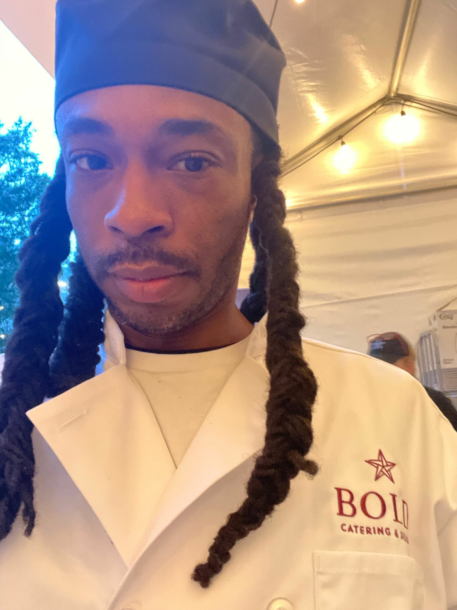 Person with long braided hair wearing a white shirt and black head covering, indoors with warm lighting.