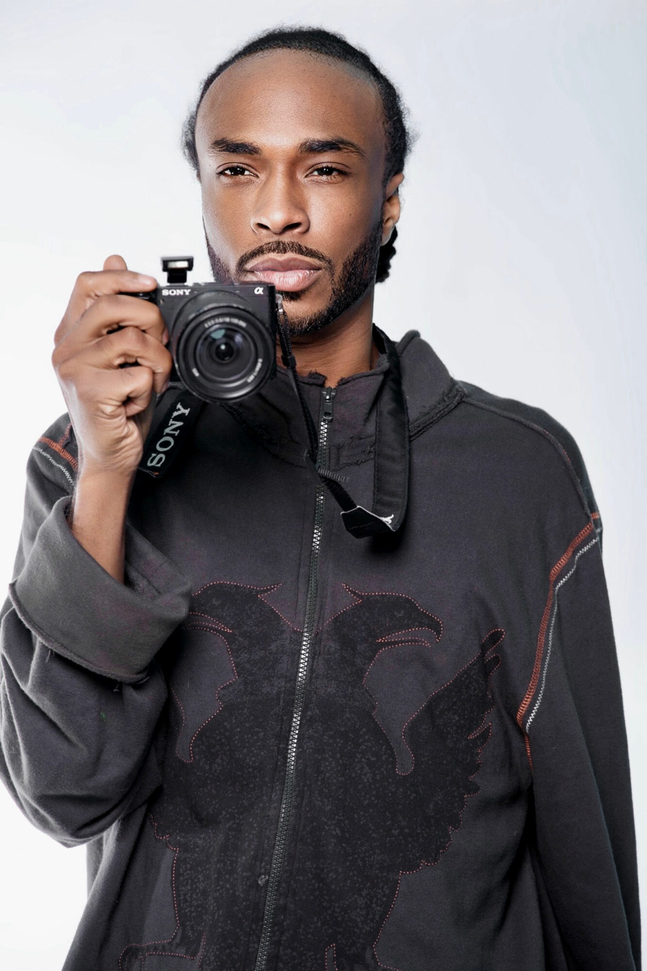 Man with a beard and braided hair holding a camera, wearing a dark hoodie, against a light background.