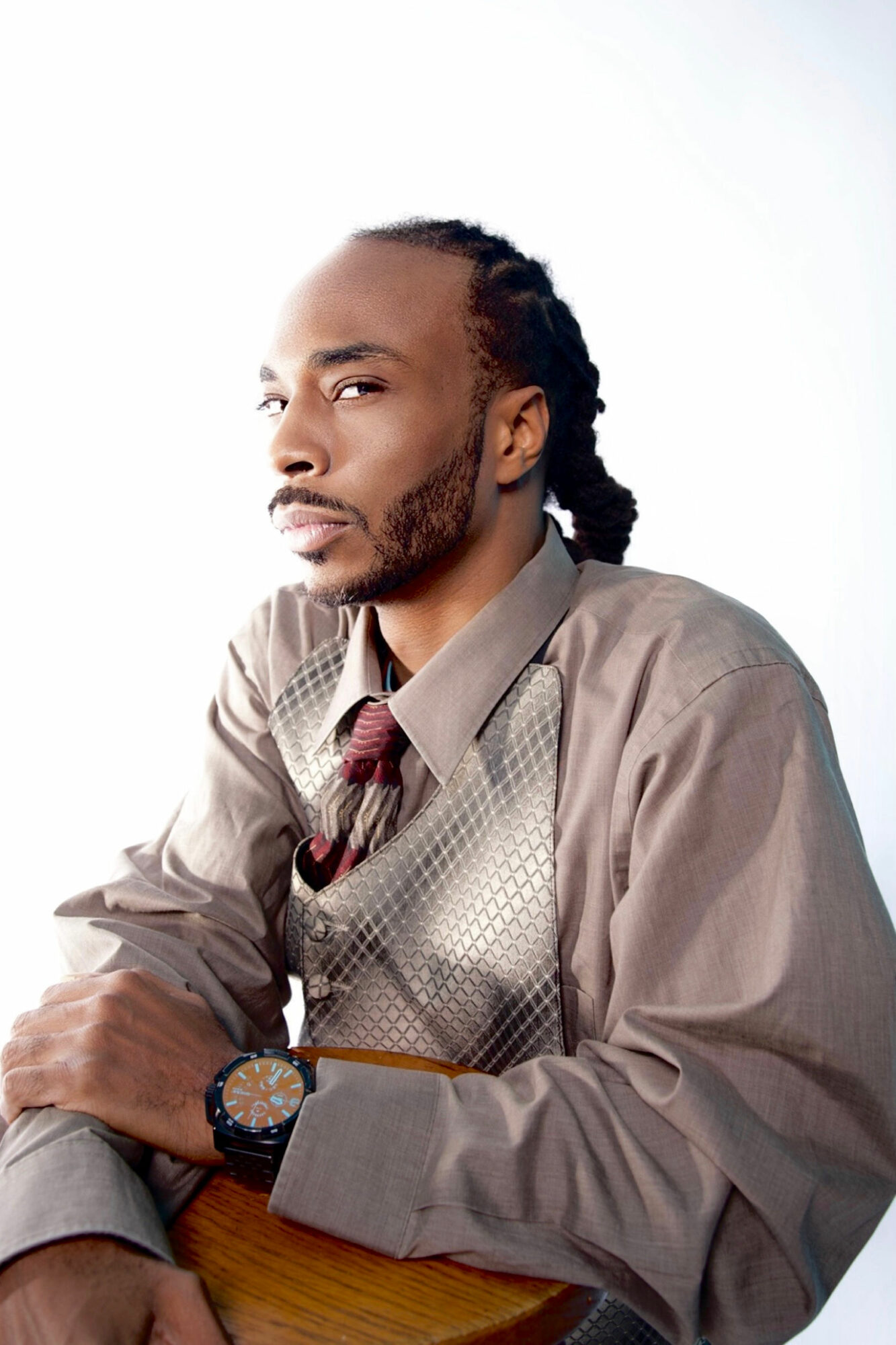 Man with braided hair and beard wearing a gray shirt and tie, sitting at a table, looking sideways.