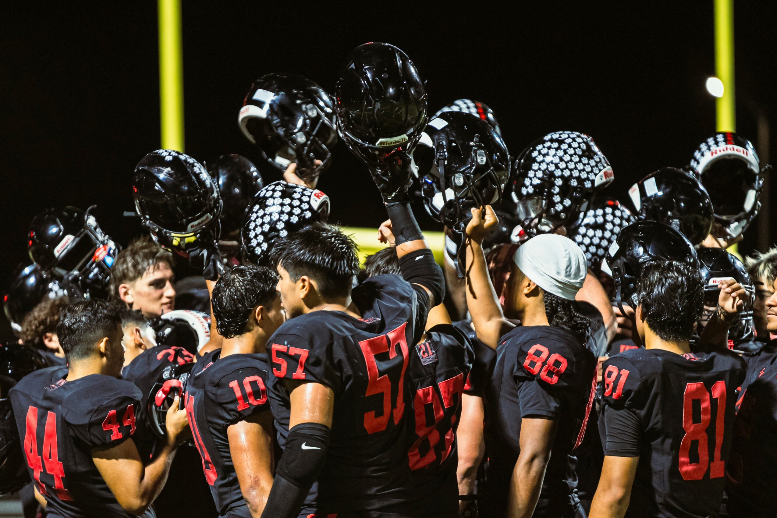 Football team in black uniforms with red numbers, gathered together with helmets raised, celebrating on field at night.