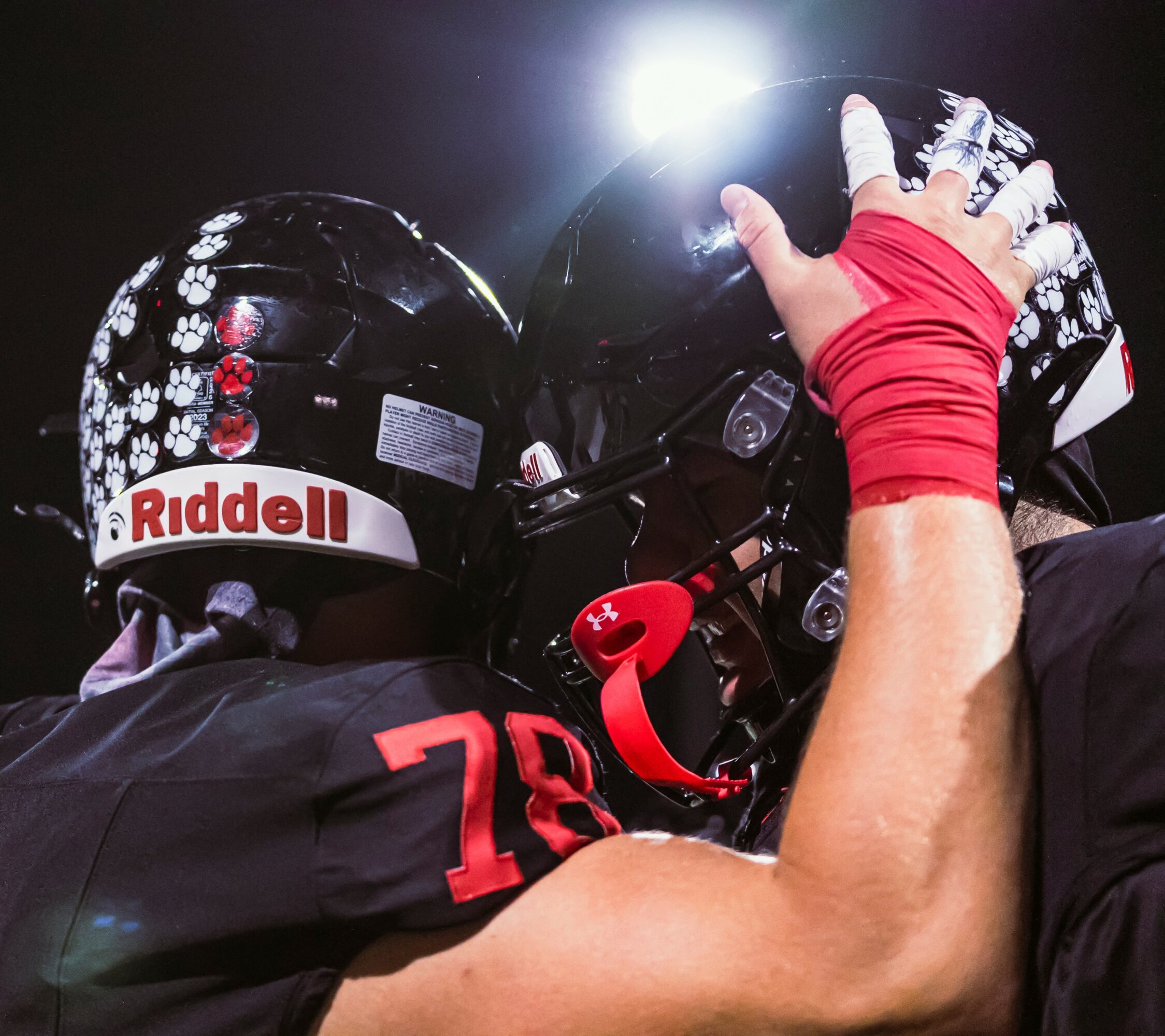 Two football players wearing helmets and uniforms, one with a red wristband, in a huddle on the field.