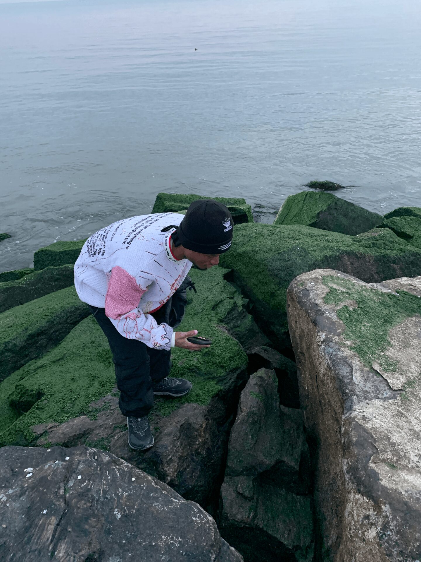 Child in white and pink jacket crouching on rocks near water, holding a phone, with green moss on rocks.