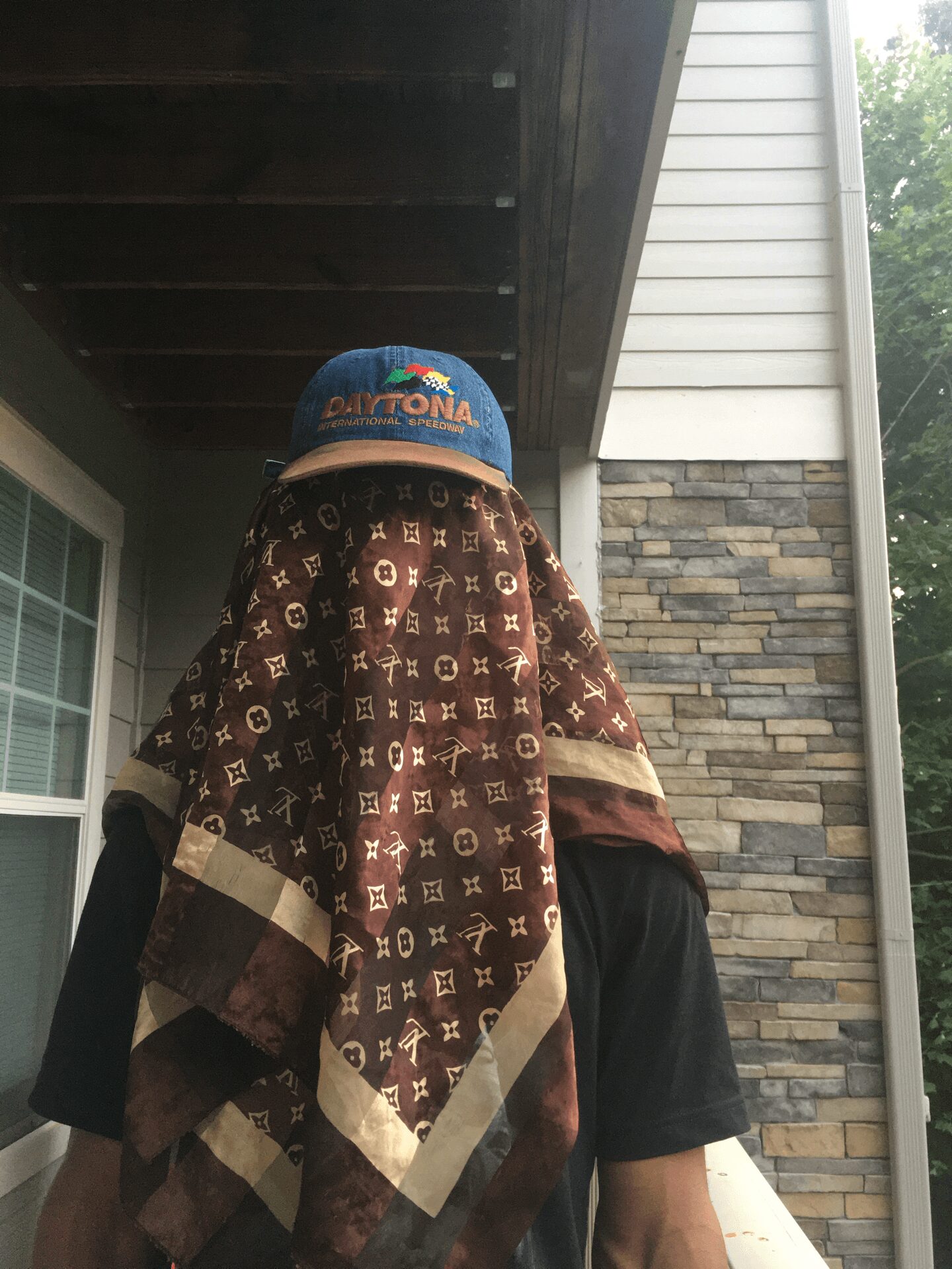 Person with a patterned cloth covering their face, wearing a blue cap, standing outside near a house with stone and siding.