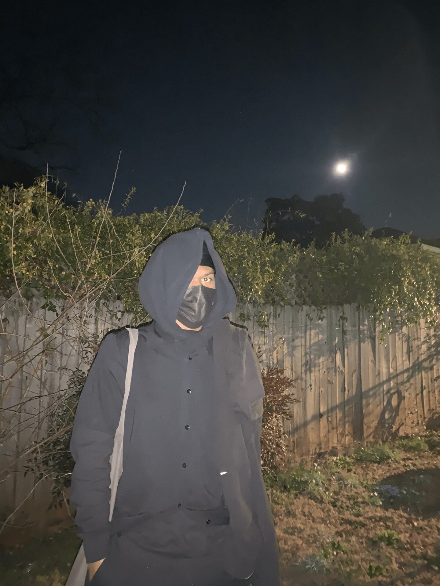 Person wearing a dark hoodie and face mask standing outdoors at night with a moonlit sky and trees in background.