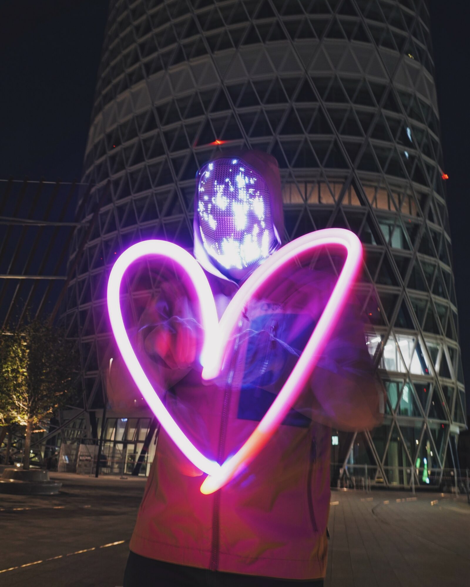 Person holding a glowing purple heart-shaped light in front of a tall building at night.