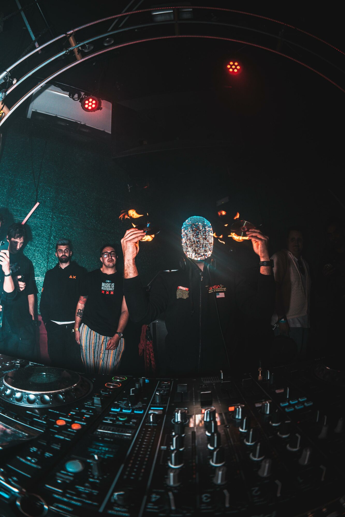Group of people on stage with DJ equipment, one person wearing a shiny mask, in a dark club setting with red lights