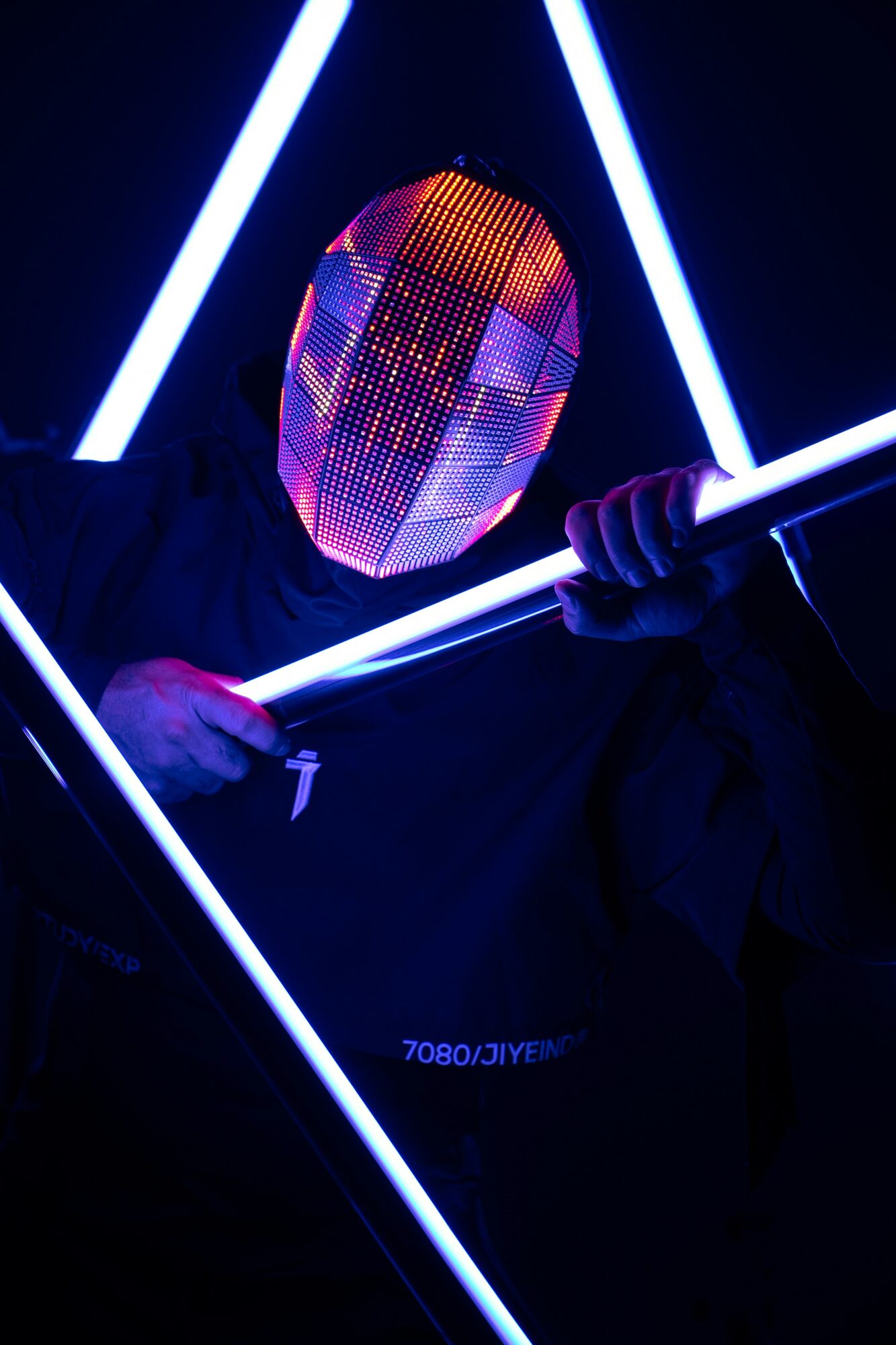Person holding glowing blue light sticks with a reflective mask, illuminated by neon lights in a dark setting.