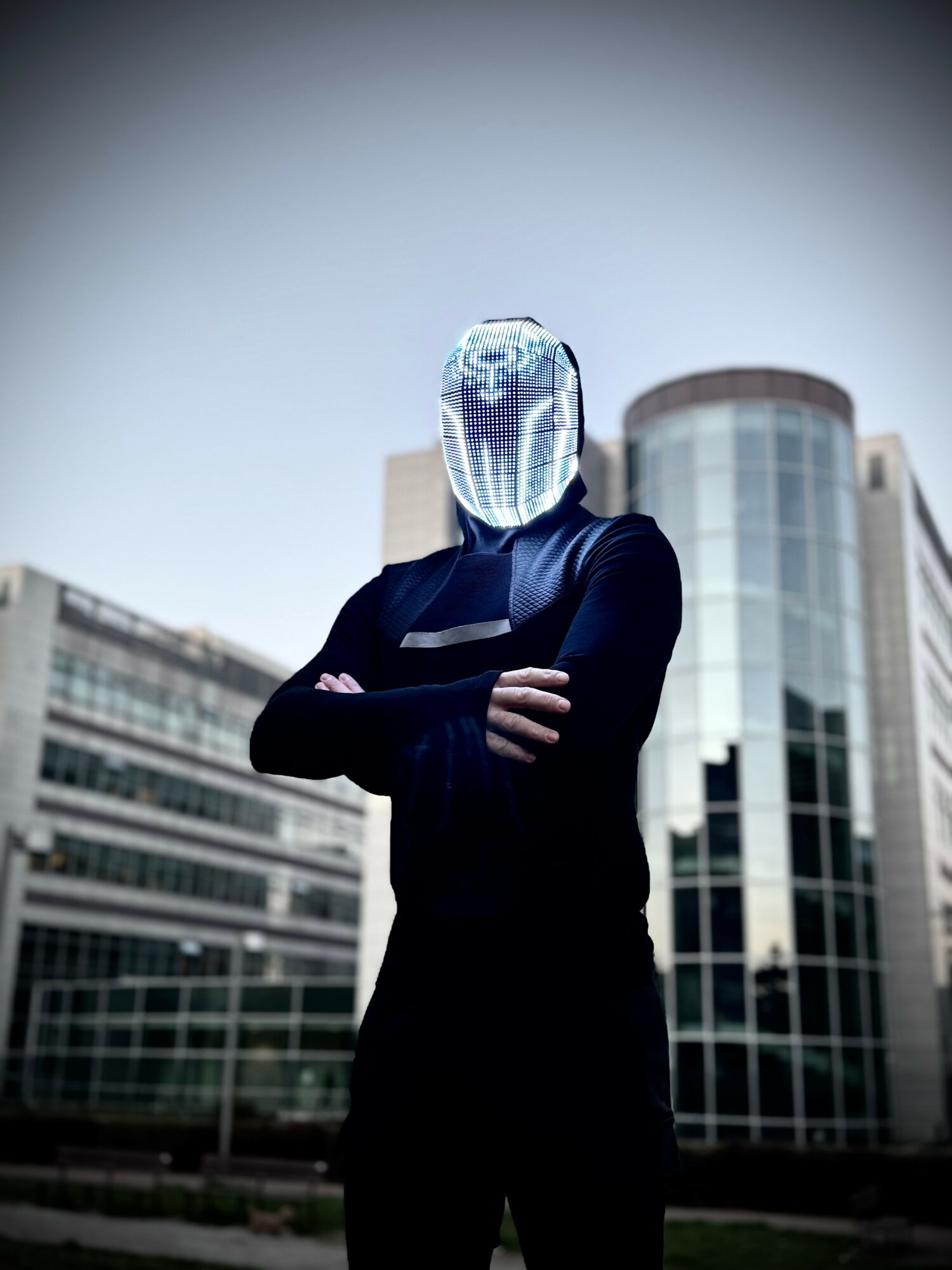 Person wearing a futuristic mask and dark clothing stands with arms crossed in front of modern office buildings.