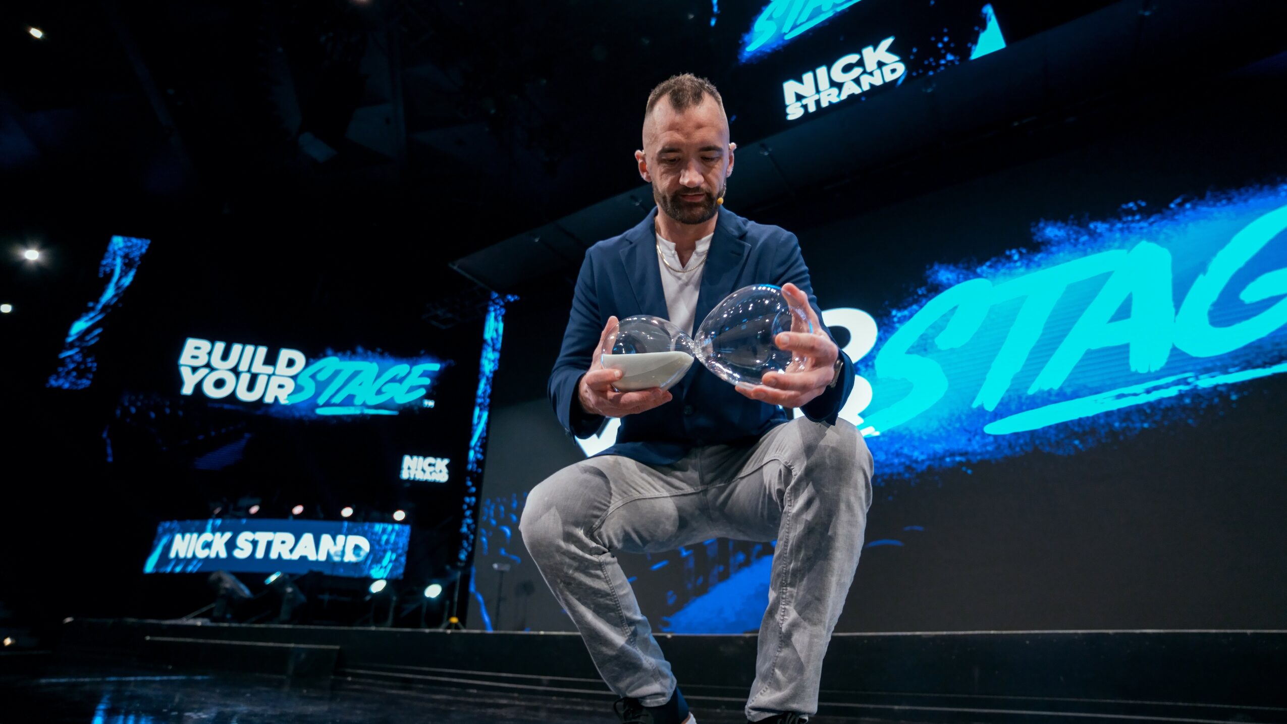 Man on stage holding an award, with screens displaying 'Build Your Stage' and 'Nick Strand' behind him.
