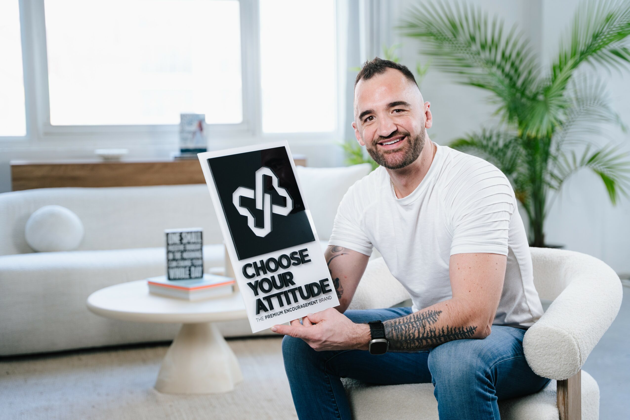 Smiling man sitting on a white chair holding a sign that says 'Choose Your Attitude' in a bright room with a large window and green plant.